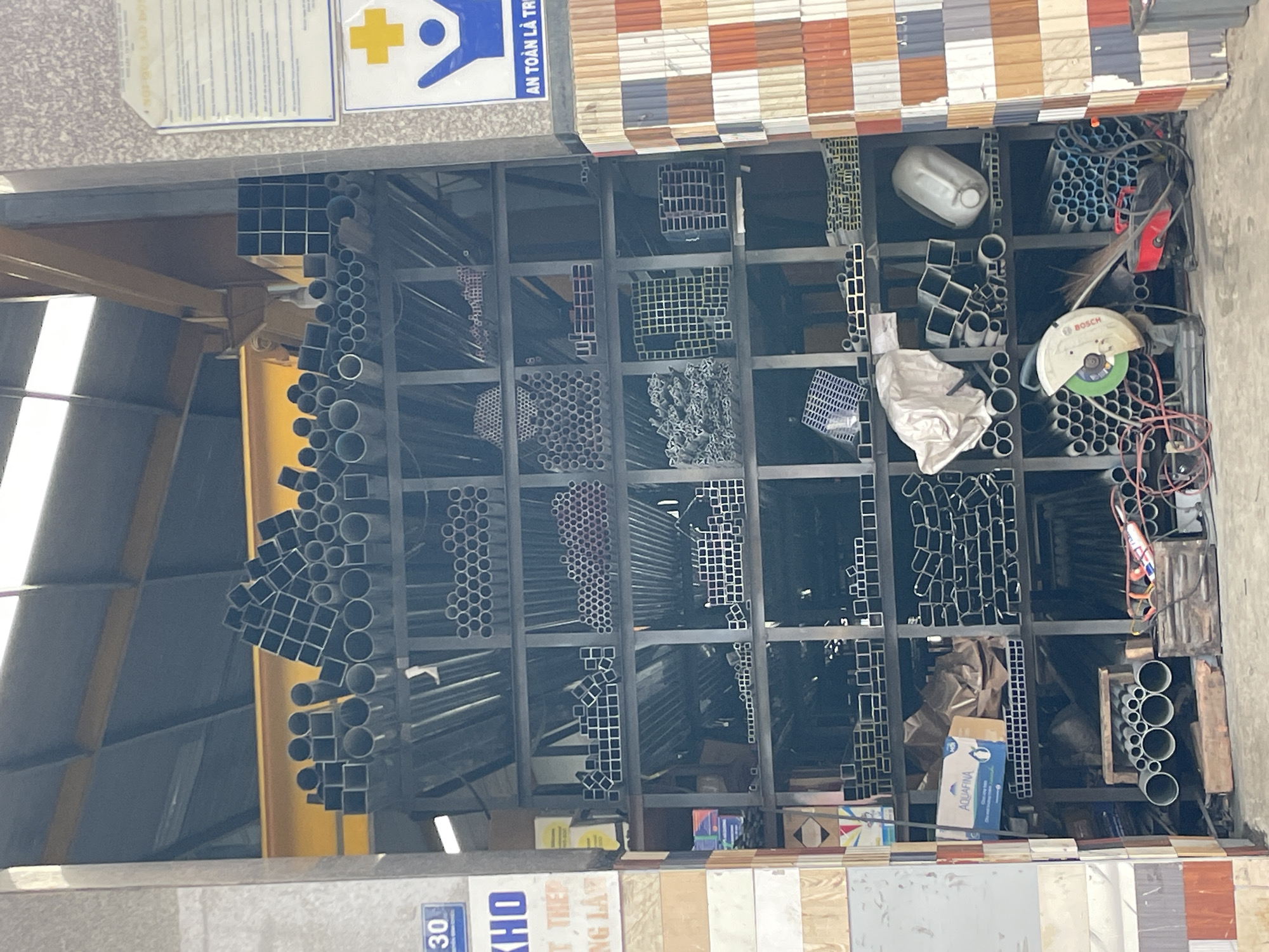 The image shows a storage area filled with metal pipes and construction materials. The shelves are packed with various sizes and shapes of metal tubing, including round and square profiles. On the left side, there are some boxes and a blue sign with white text that says "30". A sign with Vietnamese text and a yellow cross symbol is visible on the upper right. In the foreground, there is a metal cutting saw and a bundle of metal pipes. The environment appears to be a warehouse or a construction materials supplier. It is not possible to determine the specific city or country from the image alone, as there are no definitive landmarks or signs indicating Lam Vien - Da Lat, Vietnam.