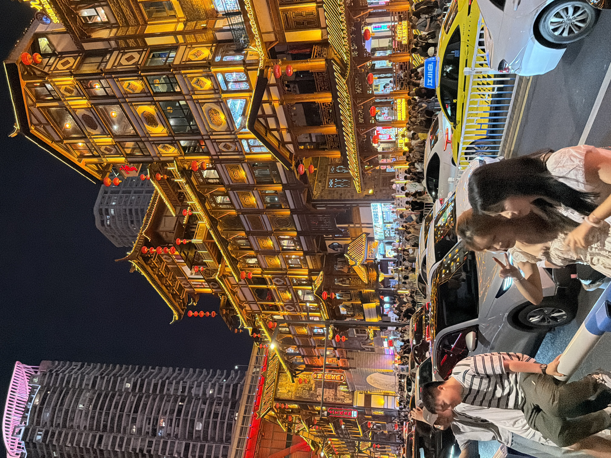 This is a nighttime, medium-shot, eye-level street scene in the Yuzhong District of Chongqing, China. The image is dominated by traditional Chinese architecture, characterized by multi-story wooden buildings with red lanterns and ornate yellow lighting. These buildings are interspersed with a modern, tall skyscraper with pink lighting on its upper floors on the left side of the frame. The street is crowded with people and vehicles, including cars and a yellow taxi with Chinese signage. In the foreground, three individuals are visible: two men and one woman. The man on the left is wearing a light-colored t-shirt and a grey baseball cap, and appears to be taking a selfie. The man in the center is wearing a black and white striped t-shirt and dark pants, and is seated on a white barrier, looking to the left. The woman on the right is wearing a light-colored dress and is gesturing with her hands, showing a peace sign. The overall impression is of a vibrant, bustling urban environment at night.