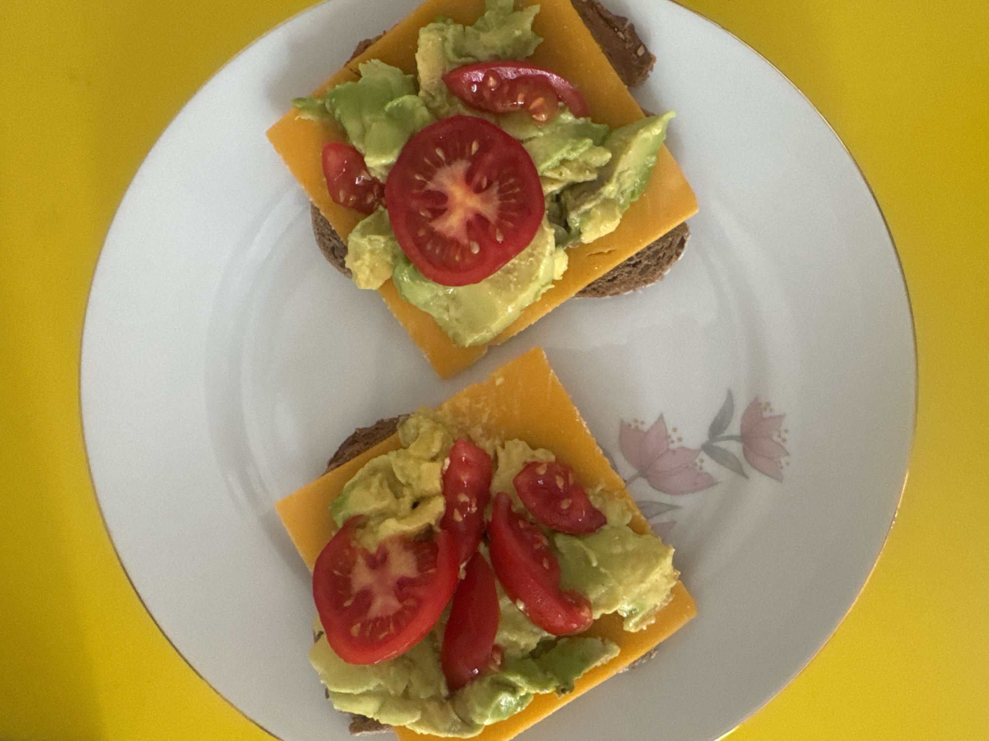 A high-angle shot depicts two open-faced sandwiches on a white plate with a floral design near the bottom. The background is a solid yellow surface. Each sandwich consists of a square slice of dark brown bread topped with a square of orange cheese, avocado chunks, and slices of red tomato. The sandwiches are positioned centrally on the plate. The plate's design features faded pink floral motifs. The scene is likely indoors, illuminated by natural light, possibly in Ramat Gan, Israel, given the user's location. No people are visible.