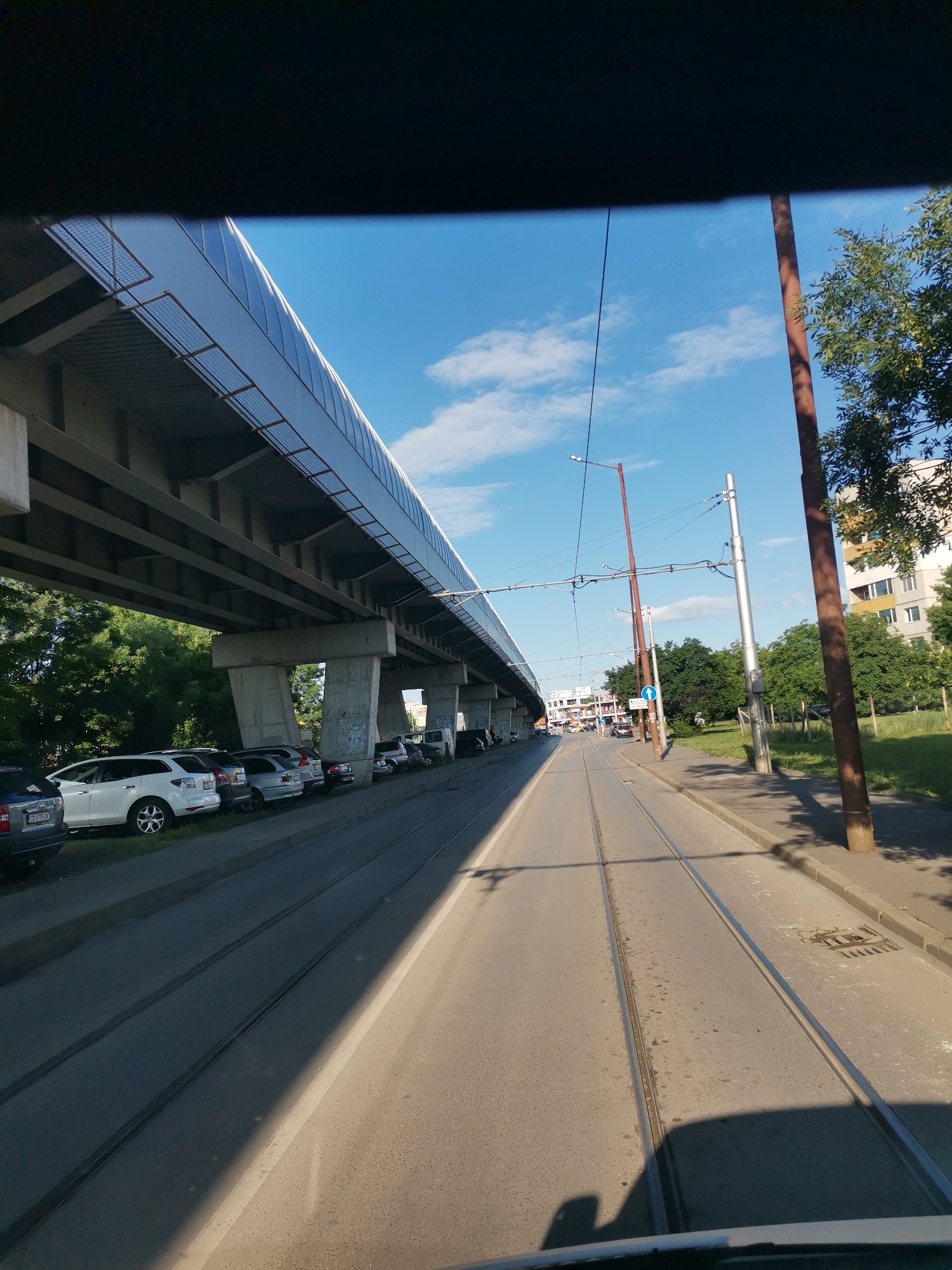 The image, taken from a moving vehicle, shows a street scene in Sofia, Bulgaria. In the foreground, tram tracks run down the center of a paved road. To the left, a multi-level elevated structure, likely a pedestrian overpass or an elevated tram line, runs parallel to the road. Beneath this structure, parked cars are visible along the side of the street, adjacent to a grassy area and trees. To the right, utility poles with overhead wires and a building are present. The sky is blue with scattered white clouds. The overall impression is of a sunny day in an urban environment with infrastructure for public transportation.