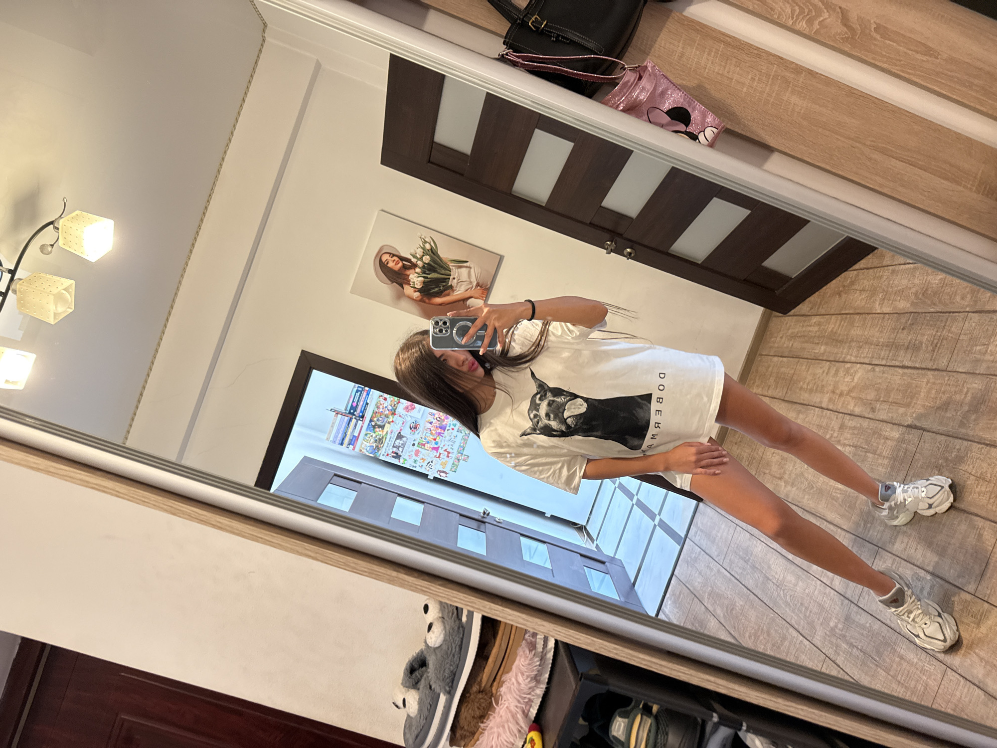 In a full-length mirror selfie taken indoors, a young woman with long, dark hair stands in a room with wooden flooring. She is wearing a white t-shirt with a black dog print and the text "DOBERMAN" printed in reverse on the bottom, white shorts, and white sneakers. She is holding a smartphone in her right hand, taking the photo. To her left, a shelf holds several pairs of slippers, including fuzzy gray ones with cartoon animal faces. Behind her, a dark brown door with frosted glass panels is visible, along with a framed picture on the wall depicting a woman holding tulips. A black handbag and a pink sequined bag hang on a rack to the right of the door. The room appears to be in a residential setting.