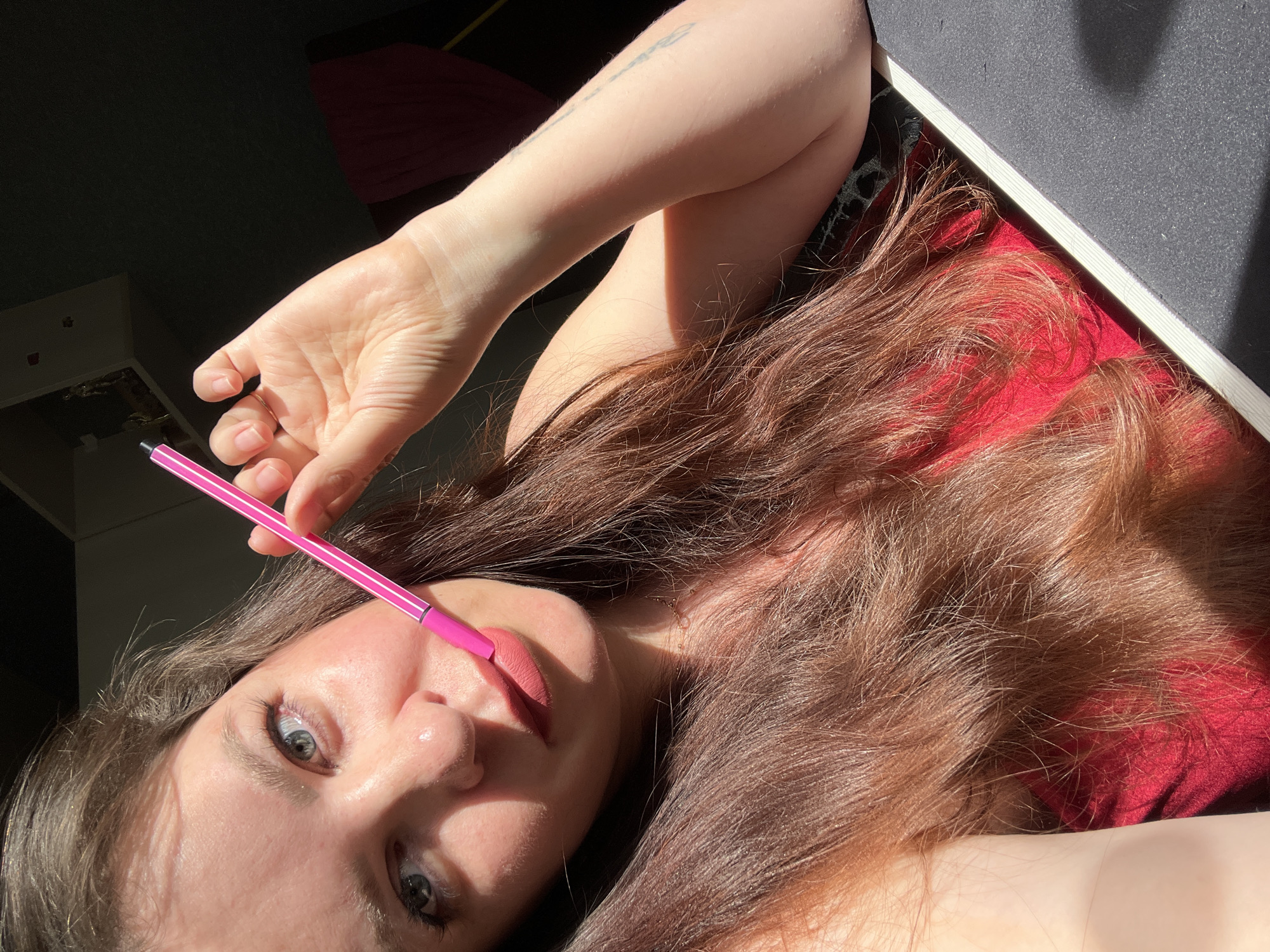 A close-up, overhead shot features a young woman lying down, with a pink marker held to her lips. She has long, wavy brown hair spread out around her and a faint tattoo on her arm. Her eyes are wide and looking directly at the camera, suggesting a playful or curious expression. The lighting indicates it is daytime, with strong sunlight casting shadows and highlighting parts of her skin and hair. The background is dimly lit, with a dark area on the left and a red fabric surface beneath her. A dark, flat object is visible on the right, with a white edge. The overall mood is intimate and candid.