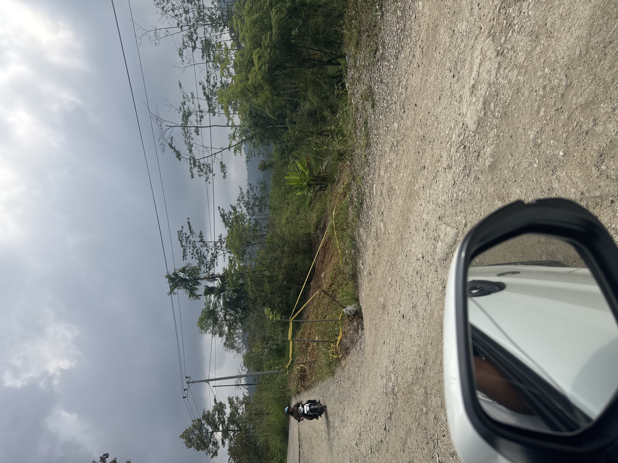 The image shows a view from inside a car looking out at a rural road in Indonesia. A person is riding a motorcycle on the dirt road. There is a yellow caution tape barrier on the side of the road, indicating a potential hazard, possibly a landslide. The landscape is hilly and covered with lush green vegetation and trees. The sky is overcast with dramatic clouds. The car's side mirror is visible in the foreground, reflecting the interior of the car.