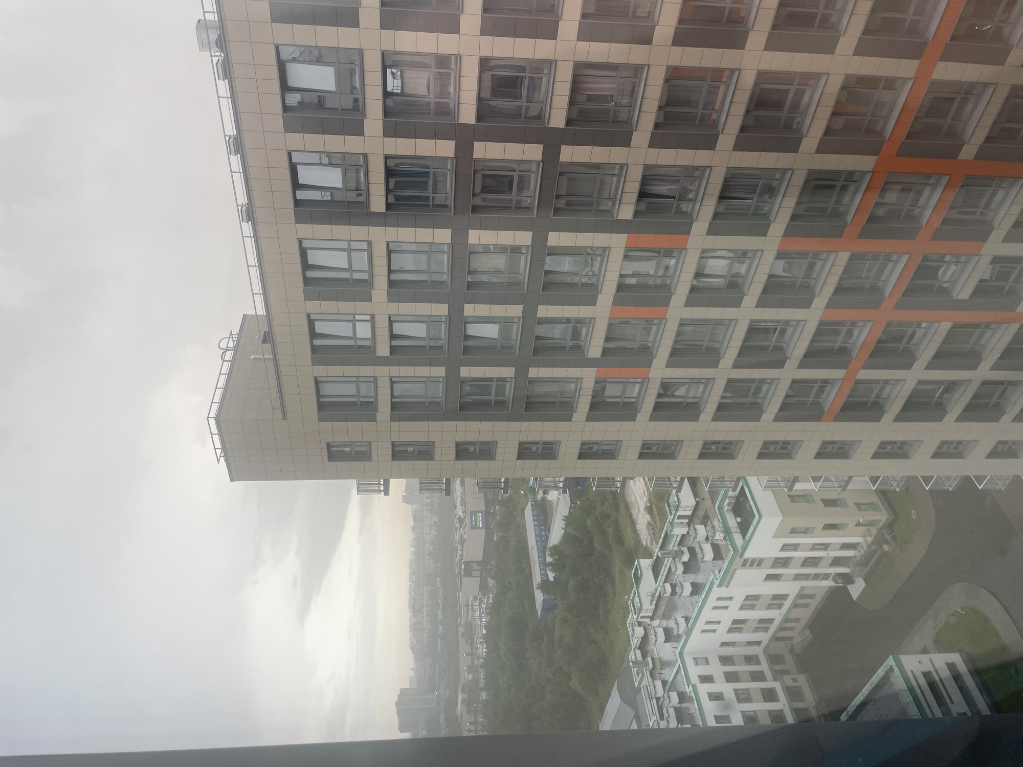 The image shows a view from a high-rise building in Poroshkino, Russia, overlooking a residential complex and distant city structures under a cloudy sky. In the foreground, a large, modern apartment building dominates the right side of the frame. Its facade is a grid of light beige and darker gray panels, with numerous windows of various sizes, some with visible curtains. Orange accent panels are arranged in a cross pattern on parts of the building. To the left and below, another residential building complex is visible. This complex is lower in height, with white and light beige sections arranged in a courtyard formation. It features multiple rooftops with ventilation units and what appear to be small balconies or protrusions. Between the two building complexes, there is a wet paved area that resembles a courtyard or driveway. Further in the distance, beyond a band of green trees, a more sprawling urban landscape unfolds with numerous taller buildings and industrial-looking structures, suggesting a city outskirt. The overall lighting is diffused and overcast, indicative of a cloudy or possibly rainy day. No people are visible in the image.