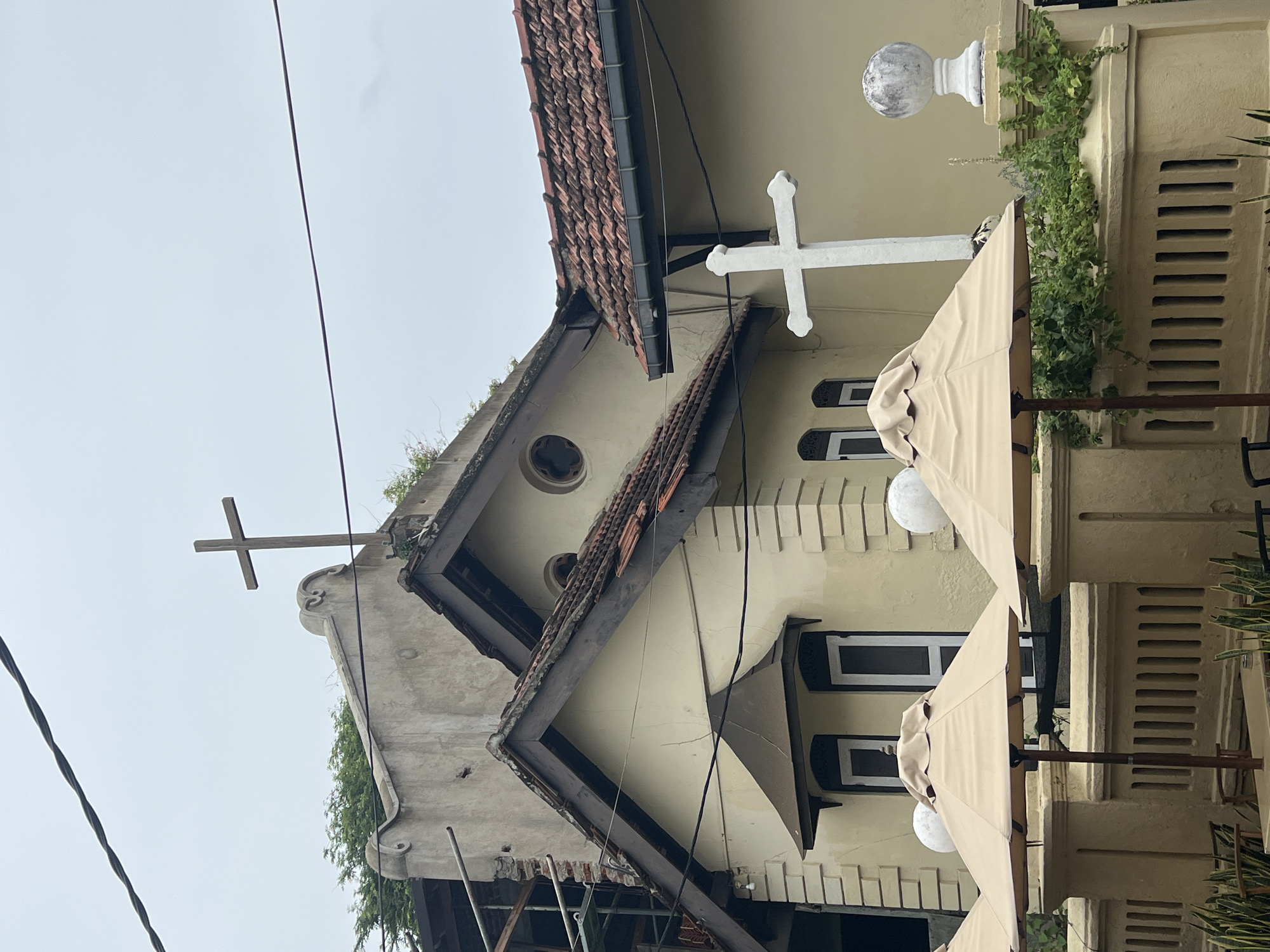 The image depicts the exterior of a colonial-era building, likely a church or a historical landmark, in Galle, Sri Lanka. The building features cream-colored walls, a red-tiled roof, and dark brown wooden trim around the gables. Two crosses are visible: a dark wooden one atop the main building and a larger white stone cross positioned in front of the structure. In the foreground, several beige patio umbrellas are set up, suggesting an outdoor seating area. A decorative, light-colored stone fence with vertical slats runs along the front of the property. Green foliage is visible growing over the fence and around the base of the white cross. The sky is overcast, indicating a cloudy day. The overall architectural style suggests a building from the Dutch colonial period.
