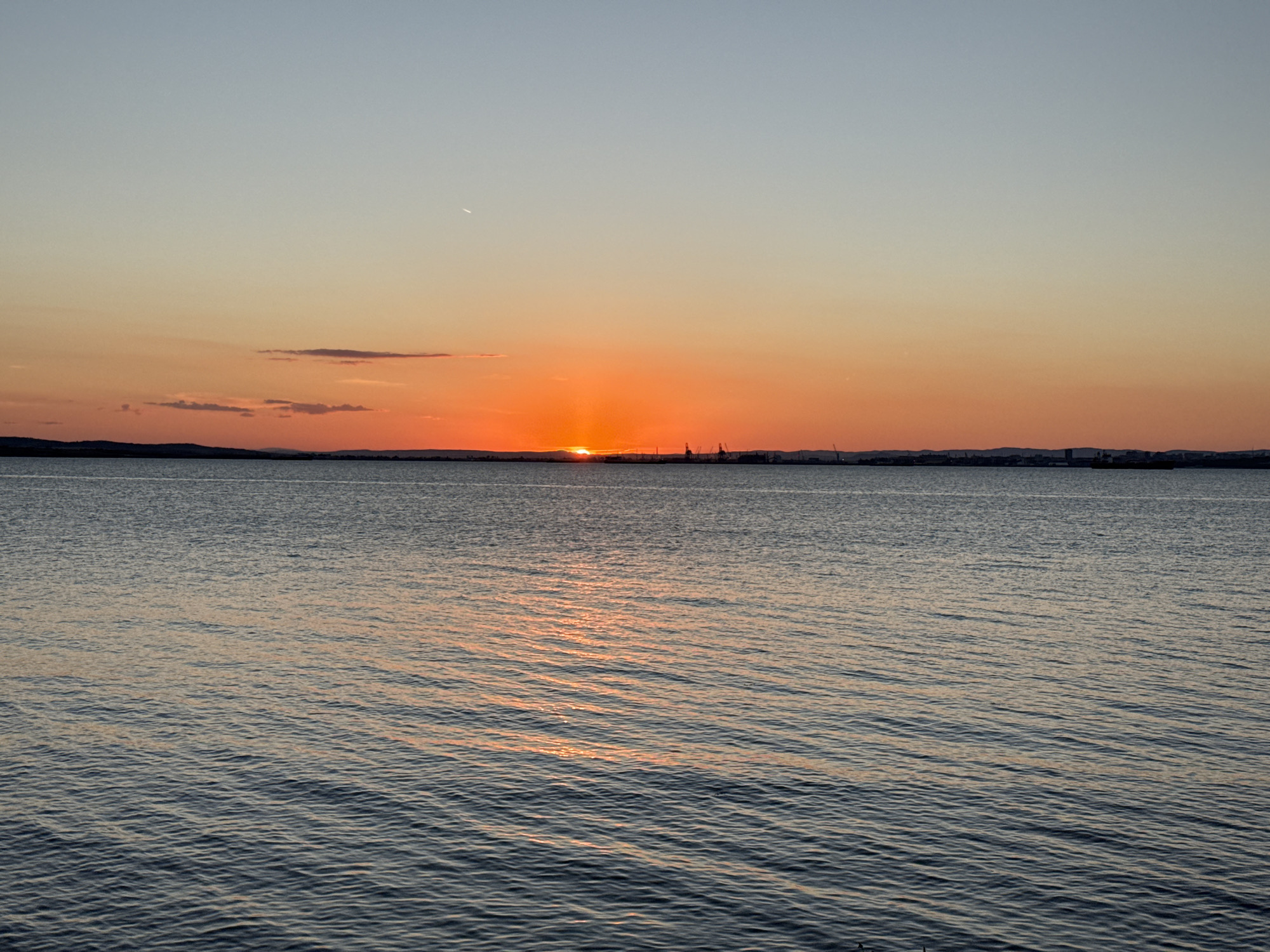 The image captures a sunset over the sea in Burgas, Bulgaria. The sun, a vibrant orange orb, is partially submerged below the horizon, casting a warm glow across the water. The sky transitions from a pale blue at the top to a gradient of orange and pink hues near the horizon, with a few wispy clouds scattered across. In the distance, the silhouette of the Burgas coastline is visible, featuring the shapes of cranes and industrial structures, hinting at the city's port. The sea is calm, with gentle ripples reflecting the colors of the sunset. The overall scene evokes a sense of tranquility and the beauty of a coastal sunset.