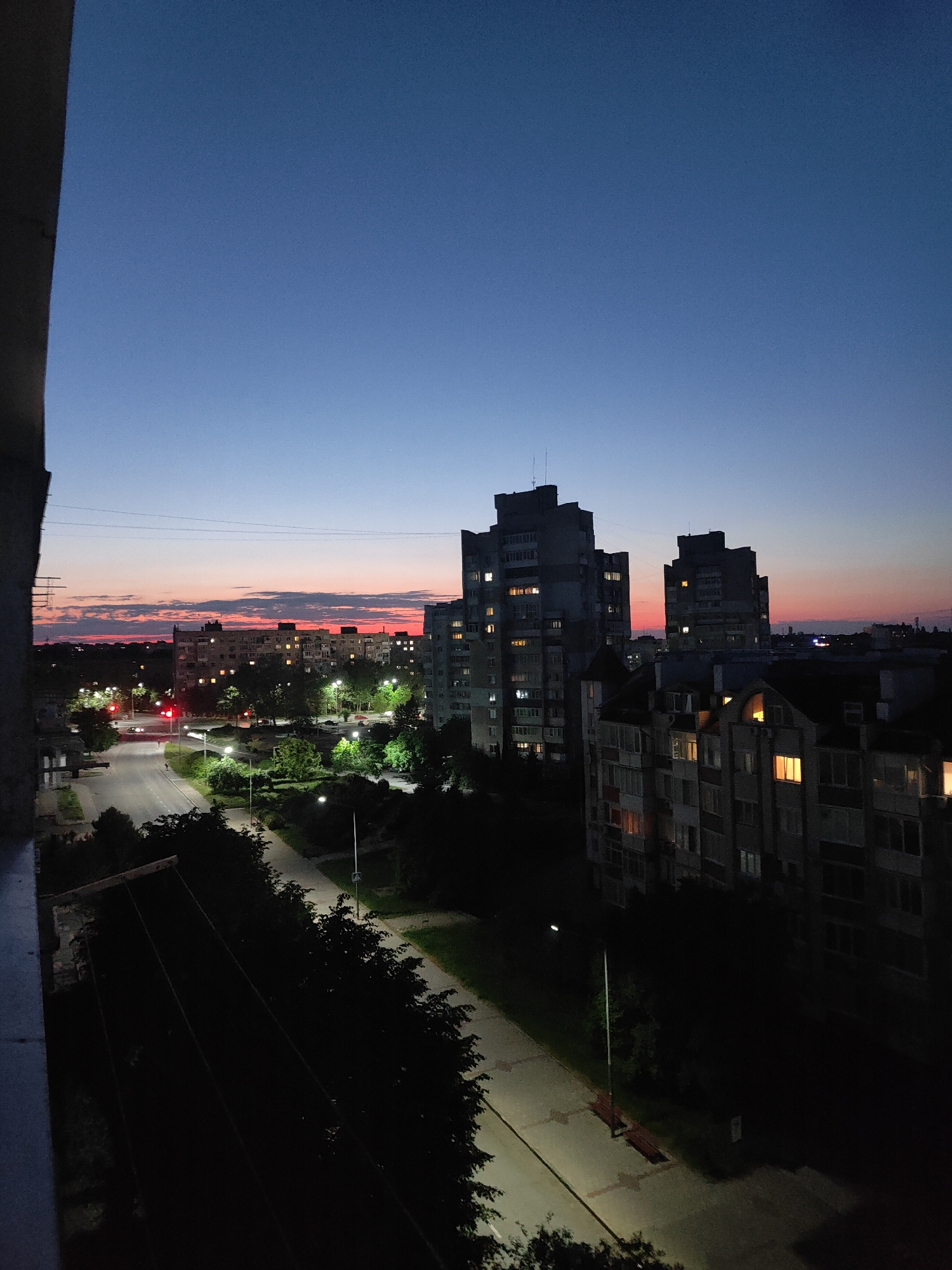 This is a vertical, high-angle photograph taken at dusk in Chornomorsk, Ukraine. The scene depicts a street and multi-story residential buildings, characteristic of a Soviet-era urban landscape. The sky is a gradient of deep blue at the top, transitioning to hues of orange and pink near the horizon, indicating the setting sun. Silhouettes of several apartment blocks dominate the middle ground and background, with lights visible in some windows, suggesting occupancy. A road runs through the lower half of the image, bordered by trees and sidewalks. Streetlights are illuminated, casting pools of light on the pavement. Power lines are visible stretched across the sky. In the foreground, the edge of a building or balcony obstructs the lower left corner. The overall mood is quiet and serene, capturing the transition from day to night in an urban environment. There are no visible people.