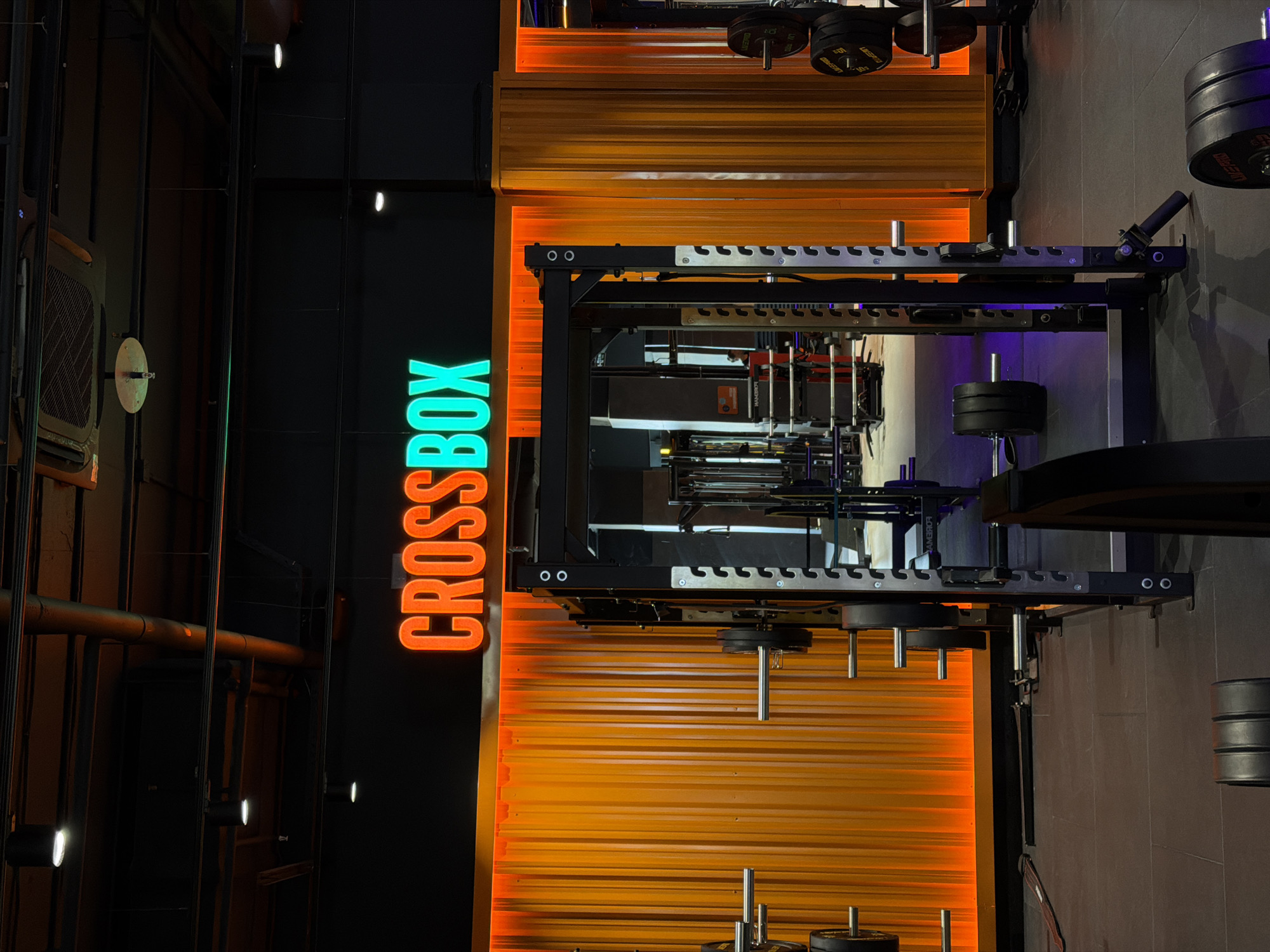 Captured indoors, the image presents a fitness facility, likely a cross-training gym, in Krasnoyarsk, Russia. The focus is on a multi-tiered training apparatus constructed from black metal, laden with weight plates. A prominent sign spells out "CROSSBOX" vertically, with orange and teal illumination. Adjacent to the training equipment, a wall displays a horizontal orange pattern. Other objects include metal pipes, ventilation units, and additional weight plates.