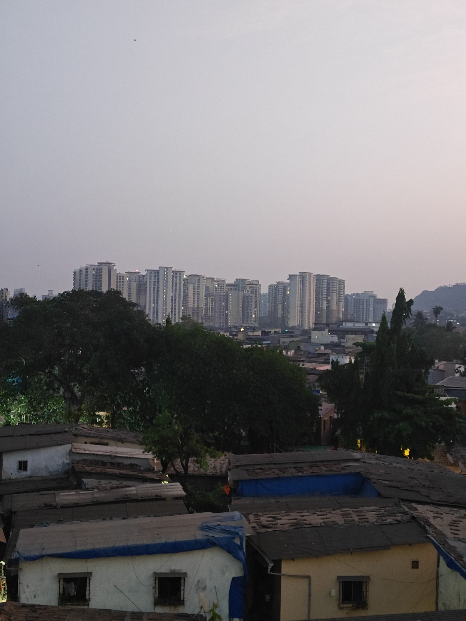 The image depicts a slum area in Mumbai, India, with low-rise buildings in the foreground and high-rise apartment buildings in the background. The sky is overcast, suggesting dusk or dawn. The slum buildings appear dense and tightly packed. Some are covered with blue tarps. Trees are scattered throughout the scene, and there is a hill visible in the distance.