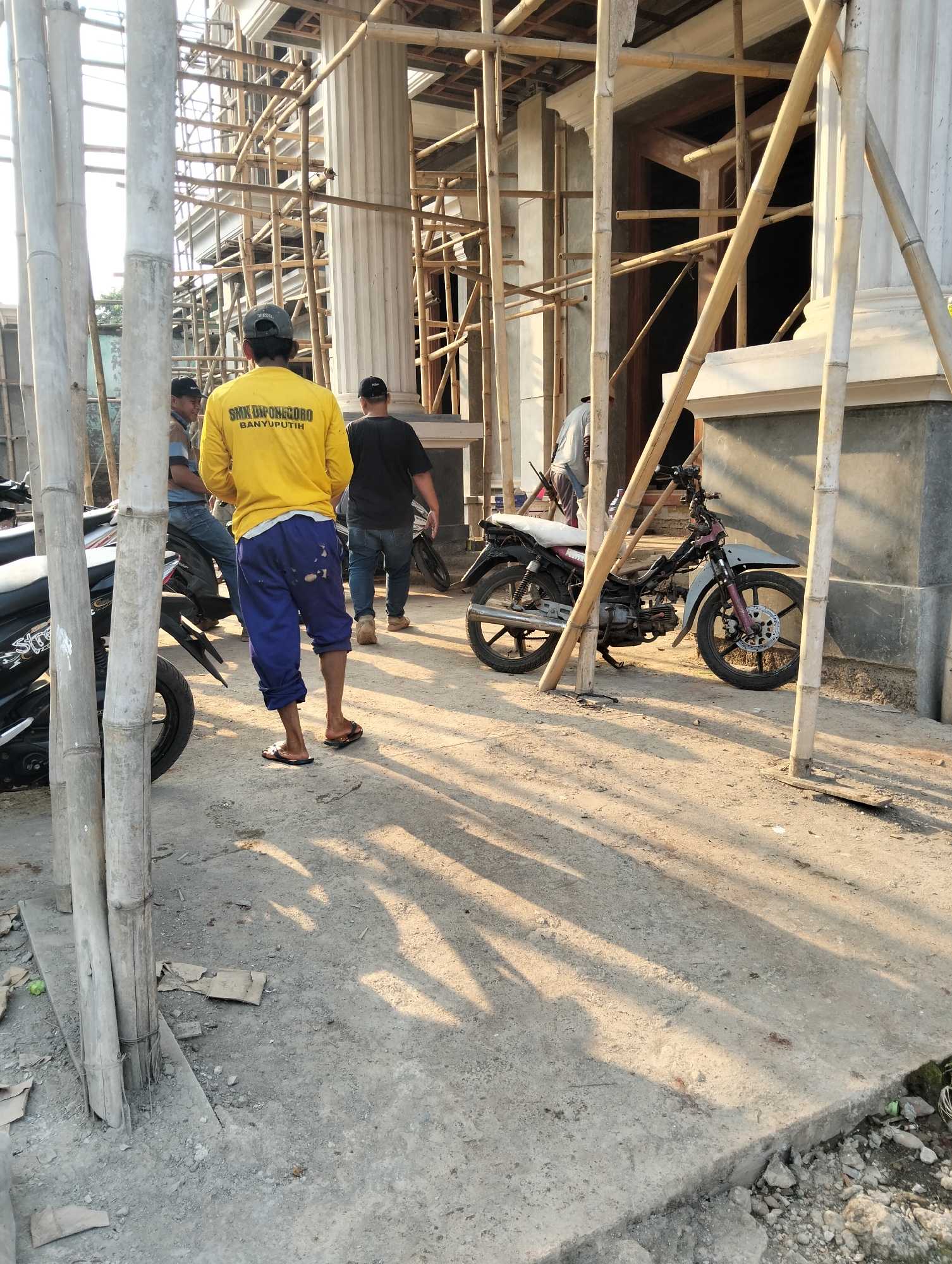 The image shows a construction site in Durenombo, Indonesia, with bamboo scaffolding surrounding a building under construction. In the foreground, three men are visible. One man in a yellow shirt with "SMK DIPONEGORO BANYUPUTIH" printed on the back is standing in the center, facing away from the camera, wearing blue pants and flip-flops. To his left, another man in a blue and brown striped shirt and jeans is sitting on a motorcycle. To the right, a man in a black shirt and jeans is walking away from the camera. A black motorcycle is parked to the right of the man in the yellow shirt, partially obscured by scaffolding. Another motorcycle is partially visible on the left side of the image. The ground is a mixture of concrete and dirt, with scattered debris. The scene is illuminated by the warm light of what appears to be late afternoon sun, casting long shadows across the ground.