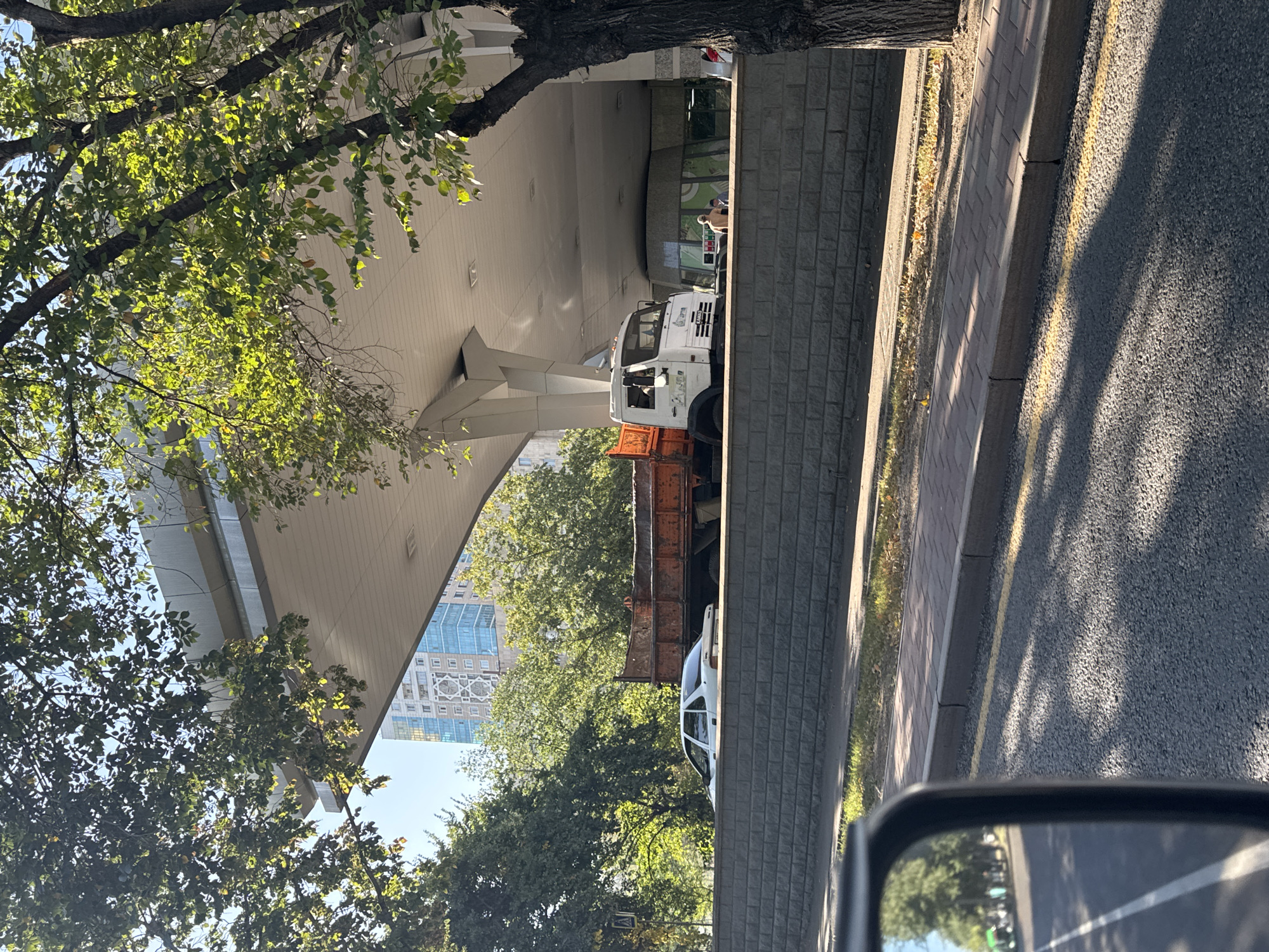 The image captures a street scene in Almaty, Kazakhstan, under a bright, sunny sky. In the foreground, a portion of a car's side mirror and the edge of the car's hood are visible, reflecting a tree-lined street with a sidewalk. The middle ground features a raised brick retaining wall separating the street from a higher level. On this upper level, a white Kamaz truck with an orange flatbed is parked. Beside the truck, a building with a curved facade and glass windows is partially visible, possibly a commercial establishment. Two individuals, one wearing a light-colored shirt and shorts, and another person are standing near the building entrance. Behind them, a tall, modern building with blue glass windows and a distinctive geometric pattern on its facade stands out against the sky, indicating an urban setting. Lush green trees with dense foliage line the area, casting shadows on the ground. The overall impression is a sunny day in a bustling city, with a mix of old and new architecture.
