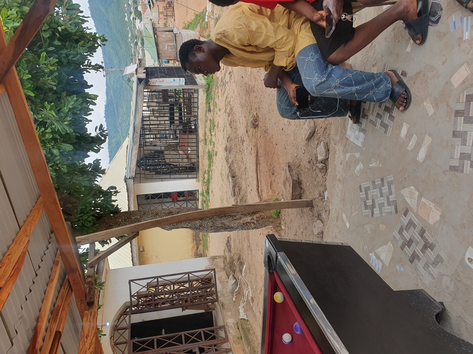 In Lokoja, Nigeria, two men are seated outdoors near a pool table. The man in the foreground, wearing a yellow shirt and blue patterned jeans, is holding a smartphone in his lap. The man to his right, partially visible, wears a red shirt and black shorts, and is also holding a smartphone. In the foreground, a red pool table with three balls (two white, one purple) is visible. The background shows a building with a corrugated metal roof supported by wooden beams, overgrown trees, and a distant view of a town nestled amongst green hills. The ground is sandy and sparsely covered with decorative tiles.