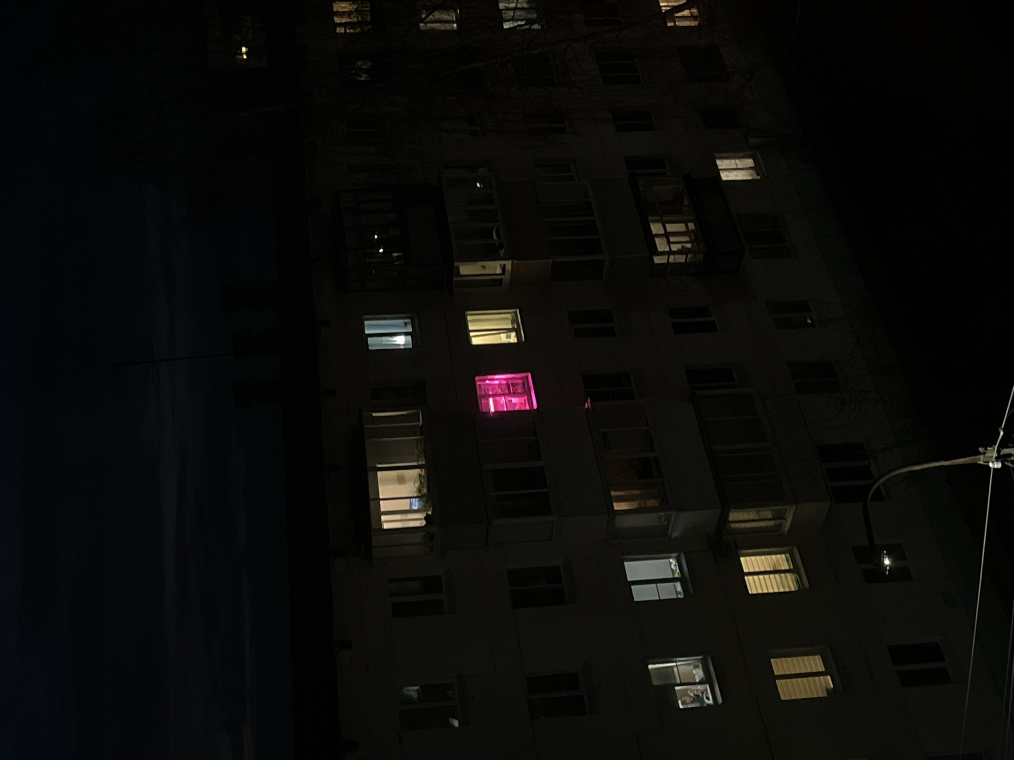 The media file captures a multi-story building at night, located in Chaikovsky, Russia. The structure, likely an apartment or residential block, is predominantly dark, with its facade rendered in deep shadows against an equally dark sky.
Various windows across different floors are illuminated, offering glimpses into the building's interior. A notable feature is a window on an upper floor that glows with a distinct, bright pink light, contrasting sharply with the warmer yellow and white lights visible in several other units. Some windows show vertical blinds, while others hint at furnishings or plants within. A few windows remain dark, blending into the building's unlit exterior.
On the far right, a segment of a utility pole or street light apparatus with overhead wires is visible, suggesting an urban or suburban setting. Faint outlines of bare tree branches can also be discerned in the upper part of the image, subtly breaking the darkness. The scene is static, capturing the quietude of nighttime with no visible human activity or text.