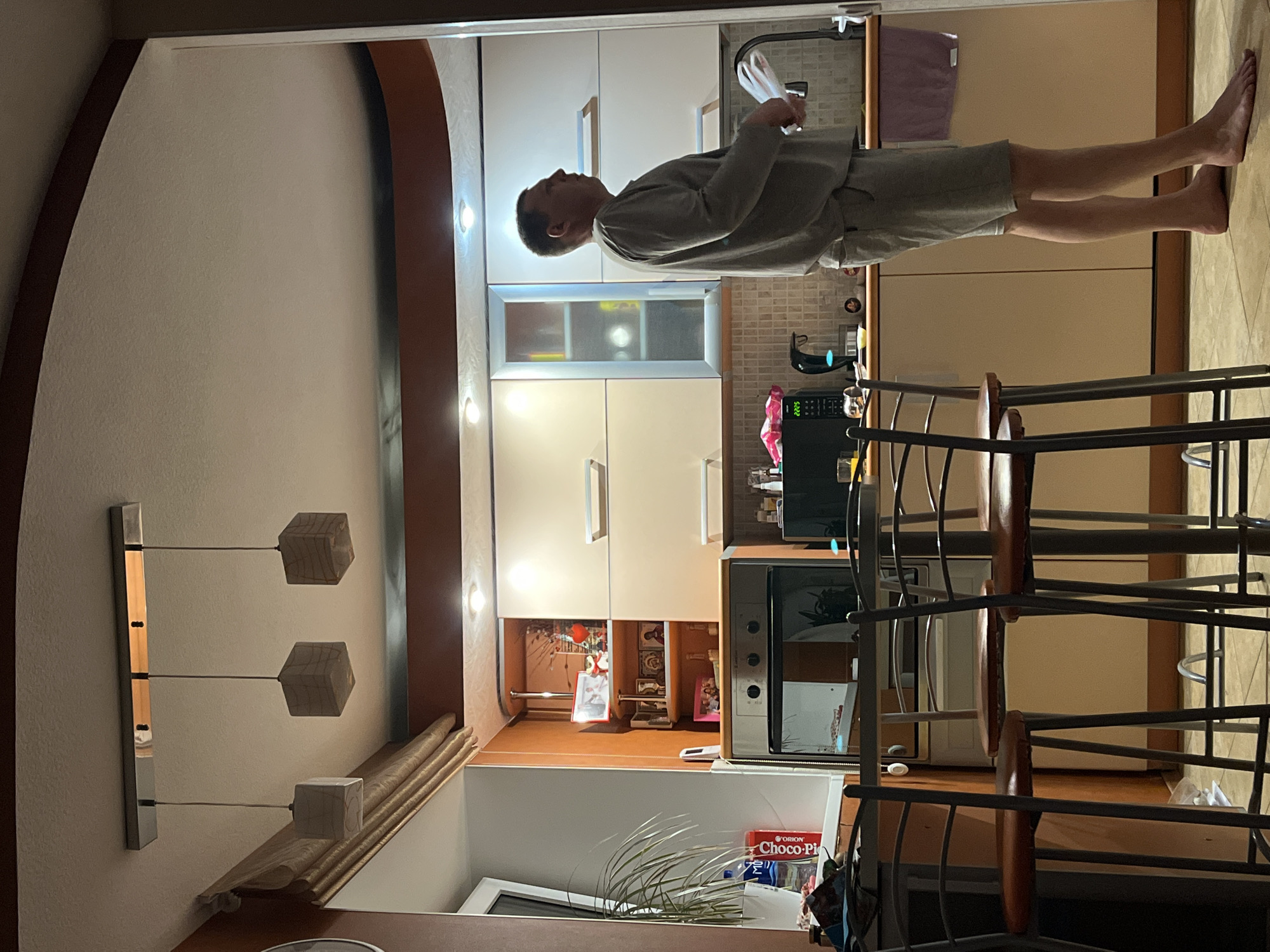 The image depicts the interior of a residential space, likely a kitchen, in Chaikovsky, Russia. A man stands in the kitchen, wearing a grey shirt and shorts, holding papers in his hands. He is positioned near a kitchen counter with cabinets and appliances, including a microwave and a coffee maker. The kitchen features a tiled backsplash, a kitchen sink, and various utensils. A bar stool stands in the foreground. The room is illuminated by overhead lighting and a modern pendant light fixture. A houseplant and a box of Choco-Pie are visible near the bottom of the image.