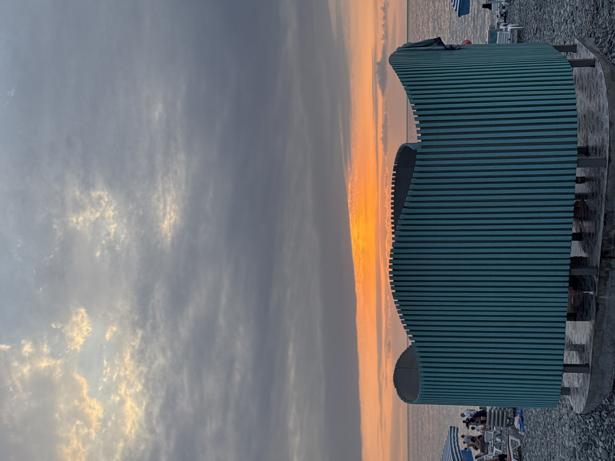 An outdoor shower stall with a teal, slatted, curved design stands on a pebble beach in Batumi, Georgia. The shower stall is centrally located in the foreground, with a concrete base. Behind the stall, a calm sea meets the horizon where a vibrant orange and yellow sunset is visible, breaking through a cloudy sky. To the left, several beach chairs and umbrellas are set up, and a group of people are gathered, some sitting and some standing. To the right, more beach chairs and a blue and white striped umbrella are visible, with the sea extending into the background. The overall atmosphere is serene, suggesting a late afternoon or early evening on the Batumi coast.