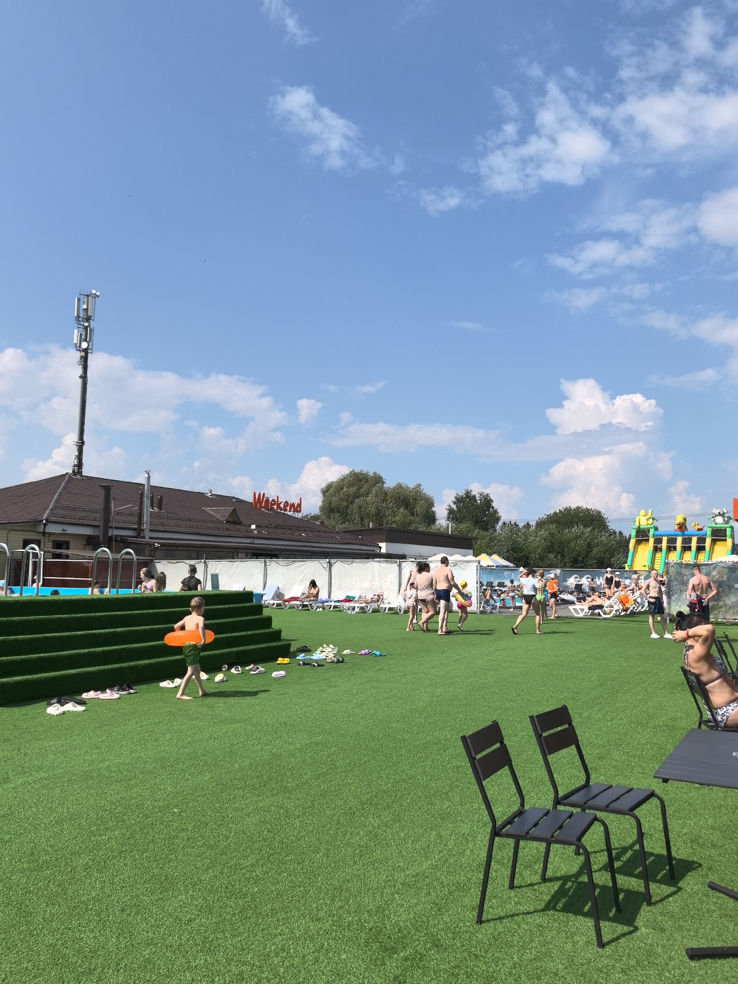 The image depicts an outdoor scene at a water park in Gamovo, Russia. The foreground features a vibrant green artificial turf area with several pairs of shoes scattered on the ground. A young boy, wearing green shorts, walks across the turf holding an orange inflatable ring. In the mid-ground, several people are visible, some walking and others relaxing on lounge chairs. A swimming pool is to the left, with people in and around it. Behind the pool, a building with a brown roof is present, featuring the red illuminated sign "Weekend" on its facade. Further in the background, a large inflatable water slide structure is set up, with people interacting with it. The sky is a clear blue, dotted with fluffy white clouds. A tall cell tower stands to the left of the frame. Two dark metal chairs and a table are placed on the artificial turf in the lower right corner.