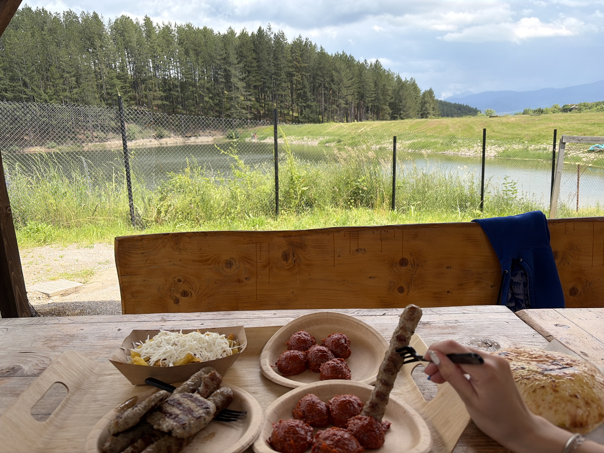 This image captures an outdoor dining scene in Alino, Bulgaria, with a natural landscape background. In the immediate foreground, a light-colored wooden table is laden with food. On the left, a paper boat contains french fries topped with a substantial quantity of shredded white cheese. Adjacent to this, a light wooden or bamboo-style plate holds several grilled meat items, including both oblong sausages (kebapche-like) and flatter patties (kyufte-like), with visible grill marks. Further right, two additional light-colored plates each contain multiple small, round, reddish-brown items, possibly a type of meatball or a spiced spread. A large, round, flat bread with a browned, slightly textured surface is positioned to the right of these plates. A human hand, with light-colored nail polish and a silver-toned bracelet on the wrist, is seen holding a black fork, piercing one of the grilled sausages.
Behind the table, a sturdy wooden bench back forms part of the seating area. A dark blue fabric item, possibly a jacket or blanket, is draped over the top edge of this bench. Beyond the dining area, a chain-link fence with dark vertical posts extends across the midground. Behind the fence lies a body of water, likely a lake or reservoir, with green vegetation along its banks. In the distance across the water, a dense forest of tall, slender coniferous trees stretches horizontally. Further beyond the trees, a wide expanse of green grassy field slopes upwards. In the far background, under a partly cloudy sky, faint blue mountain ranges are visible. Small, indistinct structures are also visible on a distant hill towards the right side of the background. Two small, indistinct figures are visible near the water's edge in the middle-left, behind the fence.