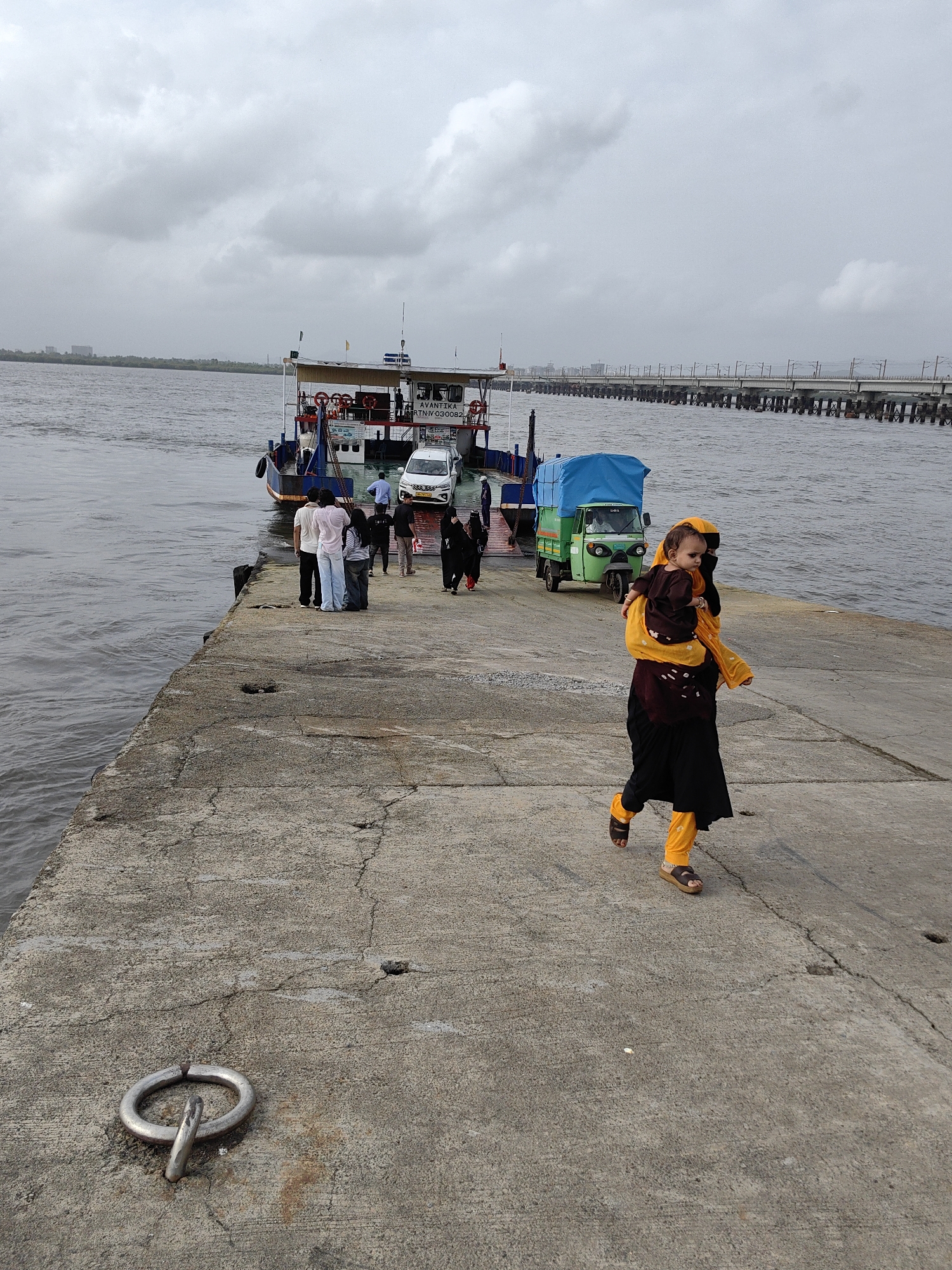 A group of people are on a concrete pier near a ferry. The pier is cracked and weathered, with a metal ring embedded in it. In the background, a ferry is docked, with vehicles and passengers visible on its deck. A green auto-rickshaw is also present on the pier. Further in the distance, a long, elevated structure, likely a bridge or railway line, stretches across the water. A woman carrying a baby on her back walks across the pier, with others standing and observing the scene. The sky is overcast, suggesting a cloudy day. The location is likely a ferry terminal in Mira-Bhayander, India, given the context of such infrastructure and transportation.