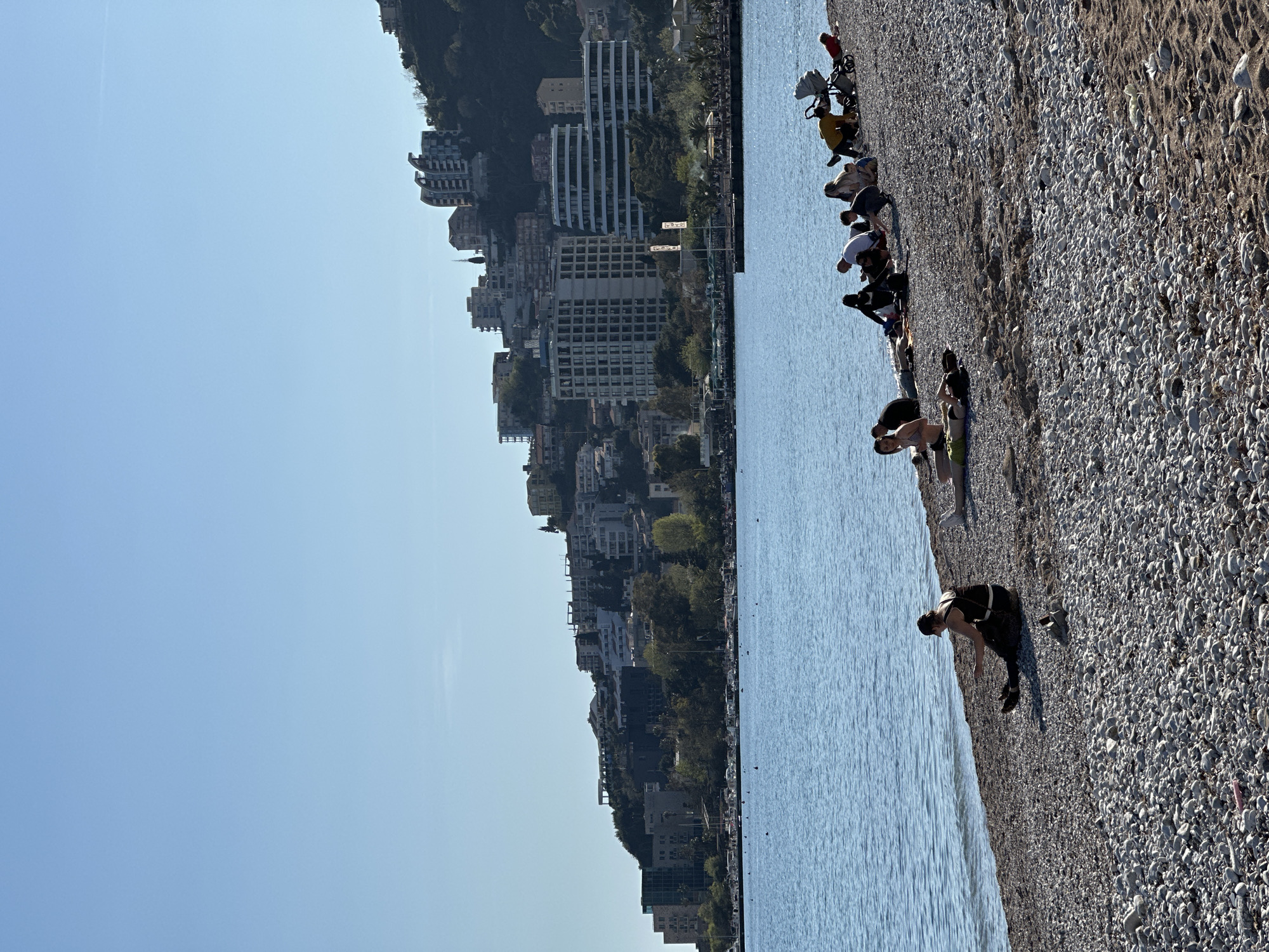 The image depicts a coastal scene likely taken in Budva, Montenegro, characterized by a pebble beach alongside a body of water, presumably the Adriatic Sea.
The beach is populated with several individuals engaging in various activities. Some are sunbathing or relaxing near the water's edge, while others are seen standing or walking along the shore. The beach material consists predominantly of small pebbles, with some sand visible in certain areas.
In the background, a cityscape is visible, featuring buildings of various heights and architectural styles. The buildings appear to be situated on a hillside, creating a tiered effect. The sky is clear and blue, suggesting a sunny day.