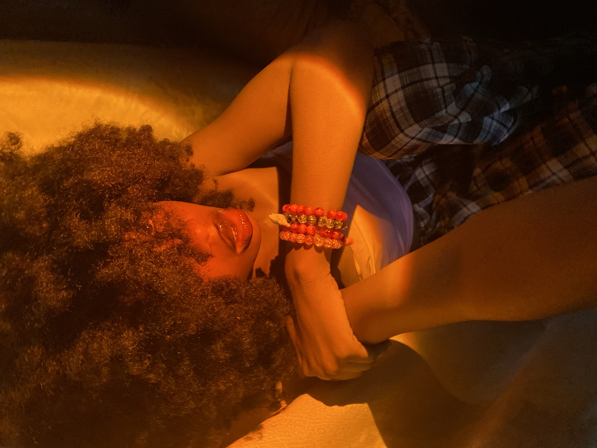 A person with brown curly hair lies on a light-colored surface. The person's tongue is visible, and they are wearing two bracelets made of orange and white beads on their left wrist. They are wearing a light purple shirt and a plaid blanket. The scene is lit by warm, orange-toned light. The location of Iranla, Nigeria is not detectable from this image alone.