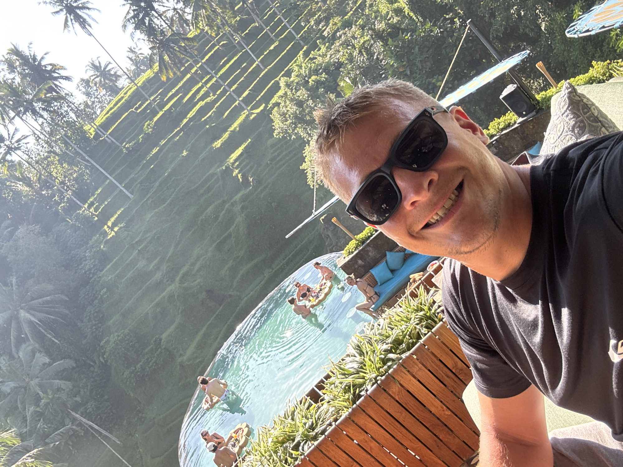 This close-up selfie captures a smiling man in the foreground, wearing black sunglasses and a black t-shirt. His face is angled slightly towards the camera, and he appears to be enjoying the sunny conditions.
Behind him, a vibrant outdoor scene unfolds. The mid-ground features an infinity swimming pool with several people relaxing in the water, some floating on inflatable rings. Around the pool area, there are comfortable-looking outdoor sofas with blue cushions and lush green plants in wooden planters.
The background reveals a magnificent landscape typical of Tegallalang, Indonesia, consisting of extensive, verdant terraced fields. Numerous tall palm trees dot the hillsides, adding to the tropical ambiance. Bright sunlight illuminates the scene, casting long shadows across the terraces, indicating a clear, sunny day, likely during the morning or late afternoon. The overall impression is one of relaxation and leisure in a beautiful natural setting. No discernible text is visible in the image.