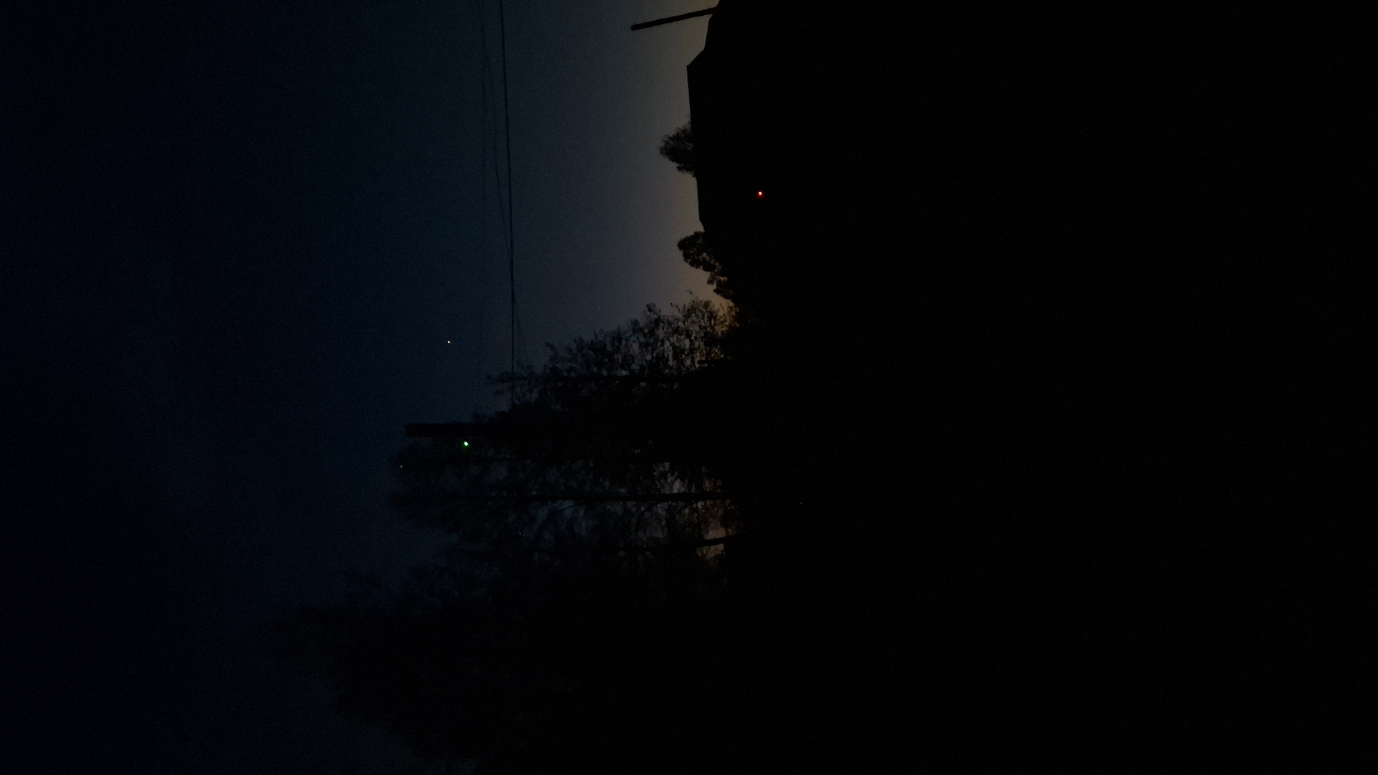 A nighttime scene in Шешурга, Russia, features a dark sky transitioning from deep blue to a lighter hue near the horizon. Silhouetted trees dominate the foreground, with a structure or building looming to the right. Two points of light are visible: a green light amidst the trees, and a single red light emanating from the structure. Power lines stretch across the frame. The scene is dominated by darkness.