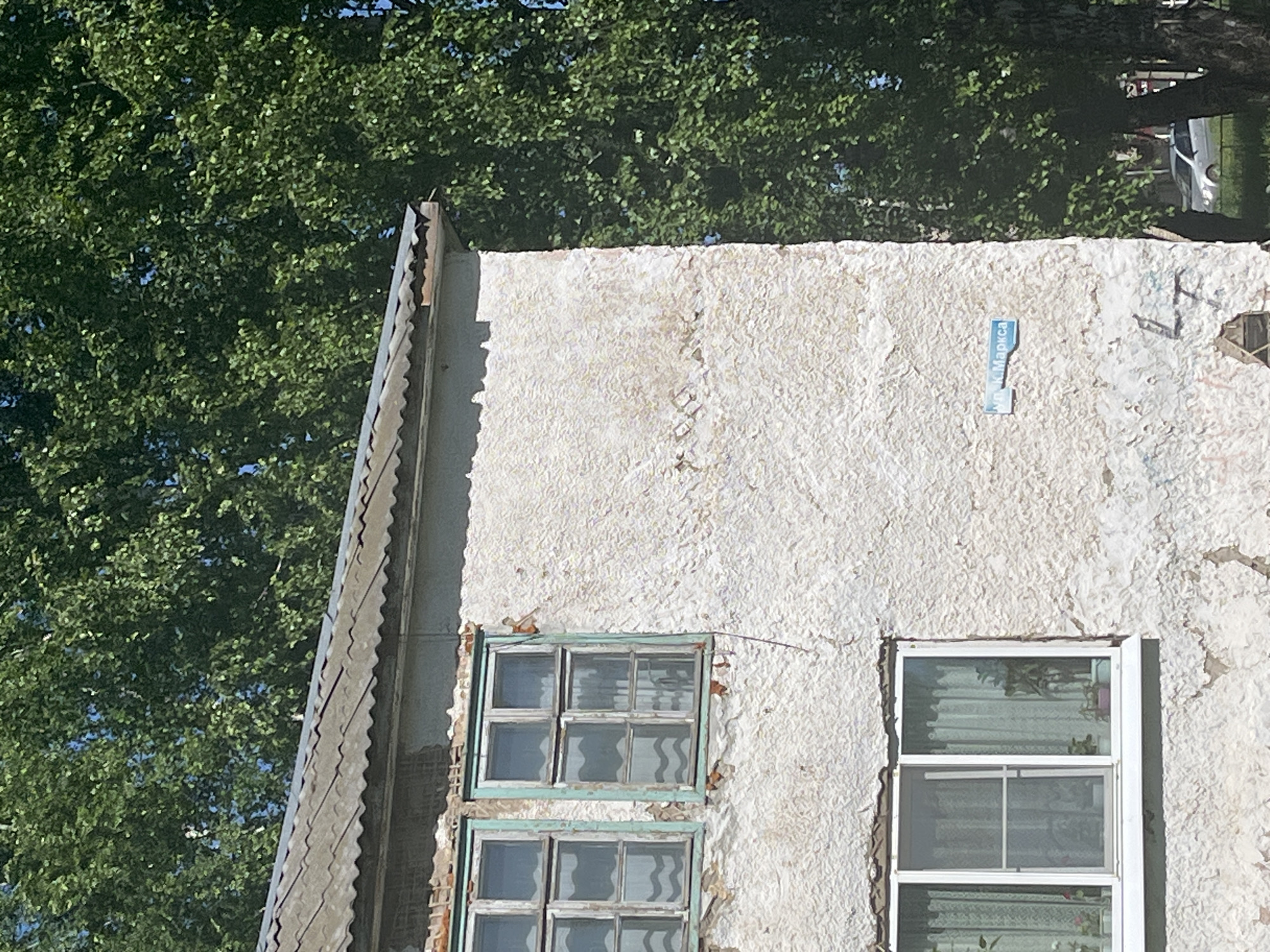 The image shows a section of an old, white-plastered building with two windows, viewed from an angle. The window on the left has a green frame and is composed of smaller panes. The window on the right has a white frame. A tree with dense green foliage is visible in the background. A street sign is affixed to the building's wall. The location is Unknown, Unknown.