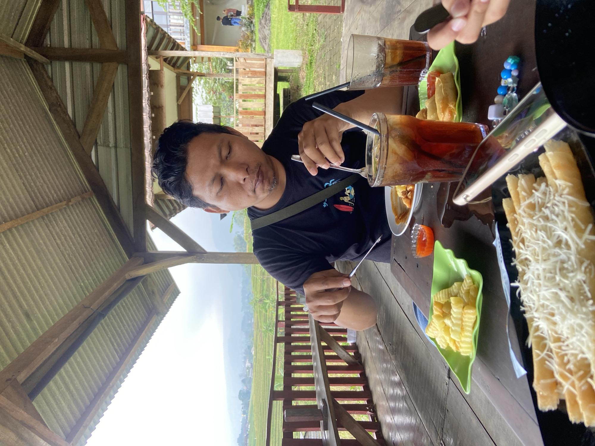 The photo shows a man sitting at a wooden table in an open-air structure with a corrugated metal roof. He wears a black t-shirt with a printed design and has a dark complexion. He is stirring a tall glass of dark liquid, likely iced tea, with a spoon. On the table, there are several plates with food, including French fries, spring rolls, and cheese sticks. Beside the plates are small dishes of what appears to be chili sauce. There are also glasses of iced tea. A smartphone is visible near the edge of the table. Outside the structure, a view of rice terraces and distant hills is visible, suggesting a rural or agricultural setting. The location is likely Penanggungan, Indonesia, judging by the type of scenery and structure shown. Other people are partially visible in the background and at the edge of the table.