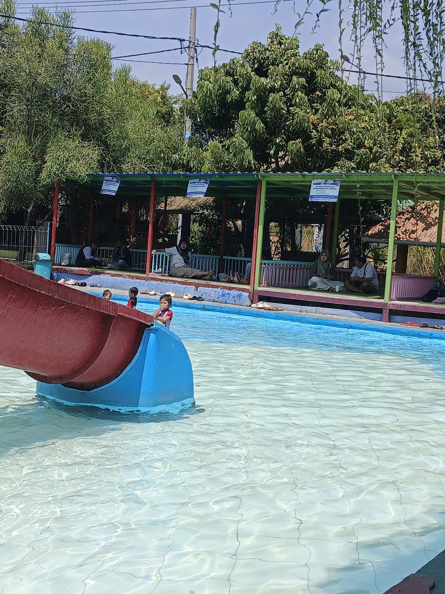 This is an outdoor medium shot image of a public swimming pool with a water slide. The pool is filled with clear, shallow water, and the bottom is visible with grid lines. A large red and blue water slide entrance is situated in the foreground, with a young child in a pink swimsuit looking towards the camera from the edge of the slide. To the left of the slide, two young boys are partially visible near the water's edge.
In the background, a covered seating area with green and red pillars is set up. Several adults are seated in this area, some are conversing while others are resting. The area is surrounded by greenery, with trees and plants visible in the background, suggesting an outdoor leisure facility. The sky is clear and blue, indicating it is a sunny day. The location is identified as Kemasantani, Indonesia.