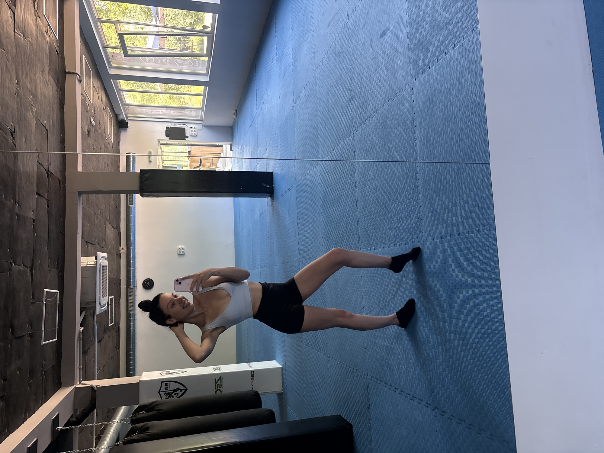 A young woman is taking a selfie in a gym in Sofia, Bulgaria. She is wearing a light blue sports bra and black shorts. She has her hair up in a bun and is smiling at the camera. The gym has blue mat flooring and punching bags in the background. The room has large windows that let in natural light.