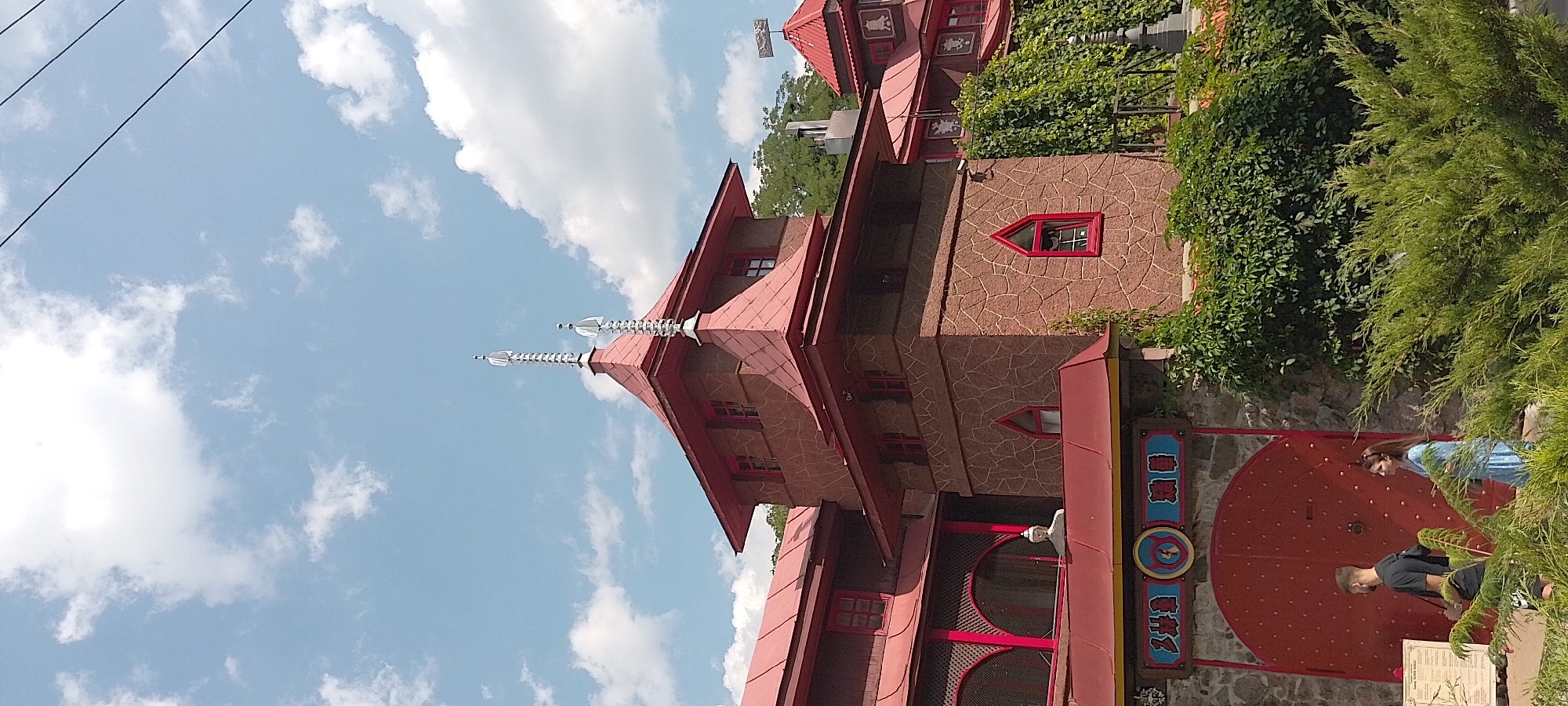 The image depicts a view of the "Hacienda" or "Dali's House" located in Cherkasy, Ukraine, an architectural landmark known for its unique, surrealist design inspired by Salvador Dalí. The building features a distinctive red tiled roof with prominent spires, a stone facade, and red-trimmed windows. In the foreground, a large, red, circular gate is visible, with two people standing near it. A man in a black t-shirt and shorts is on the left, and a woman in a blue dress is on the right. Lush green bushes and trees surround the lower part of the building, with ivy growing up the stone walls. The sky above is blue with scattered white clouds, and electrical wires are visible stretching across the upper portion of the frame. The overall scene captures the unconventional architecture of the building set against a natural environment on a clear day.