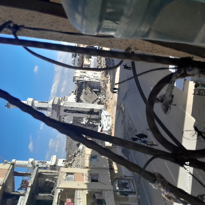 An elevated viewpoint captures a street scene in Khan Yunis, Palestinian Territory, under a clear blue sky. The foreground is partially obscured by a dark, metallic grate or railing, with a large, translucent blue-green water bottle visible on the right.
The midground features a paved road running horizontally, with an intersection or curve visible. To the right of the road, a severely damaged mosque stands prominently, characterized by a partially standing minaret and a second minaret further right. The main structure of the mosque is largely collapsed, with its roof missing and significant sections of walls destroyed, revealing internal debris and rubble. White tarpaulins or makeshift shelters are visible near the mosque's base.
To the left of the road, a multi-story building exhibits extensive damage, including missing facade sections on multiple floors, exposing interior spaces. Laundry hangs from a balcony on an upper, less damaged floor. The ground level of this building shows partial structural integrity.
On the street, approximately six to eight individuals are present. Two dark-clad figures are walking amidst debris near the damaged mosque on the right side of the road. A group of three to four individuals, including adults and possibly children, walk together on the left side of the road, heading away from the viewer. A lone figure on a bicycle or scooter moves further down the street on the right. A small figure is visible in the lower right corner, near a ladder-like object. The surrounding areas on either side of the road are strewn with rubble and dust, remnants of destroyed structures. The scene is illuminated by strong daylight, casting visible shadows.