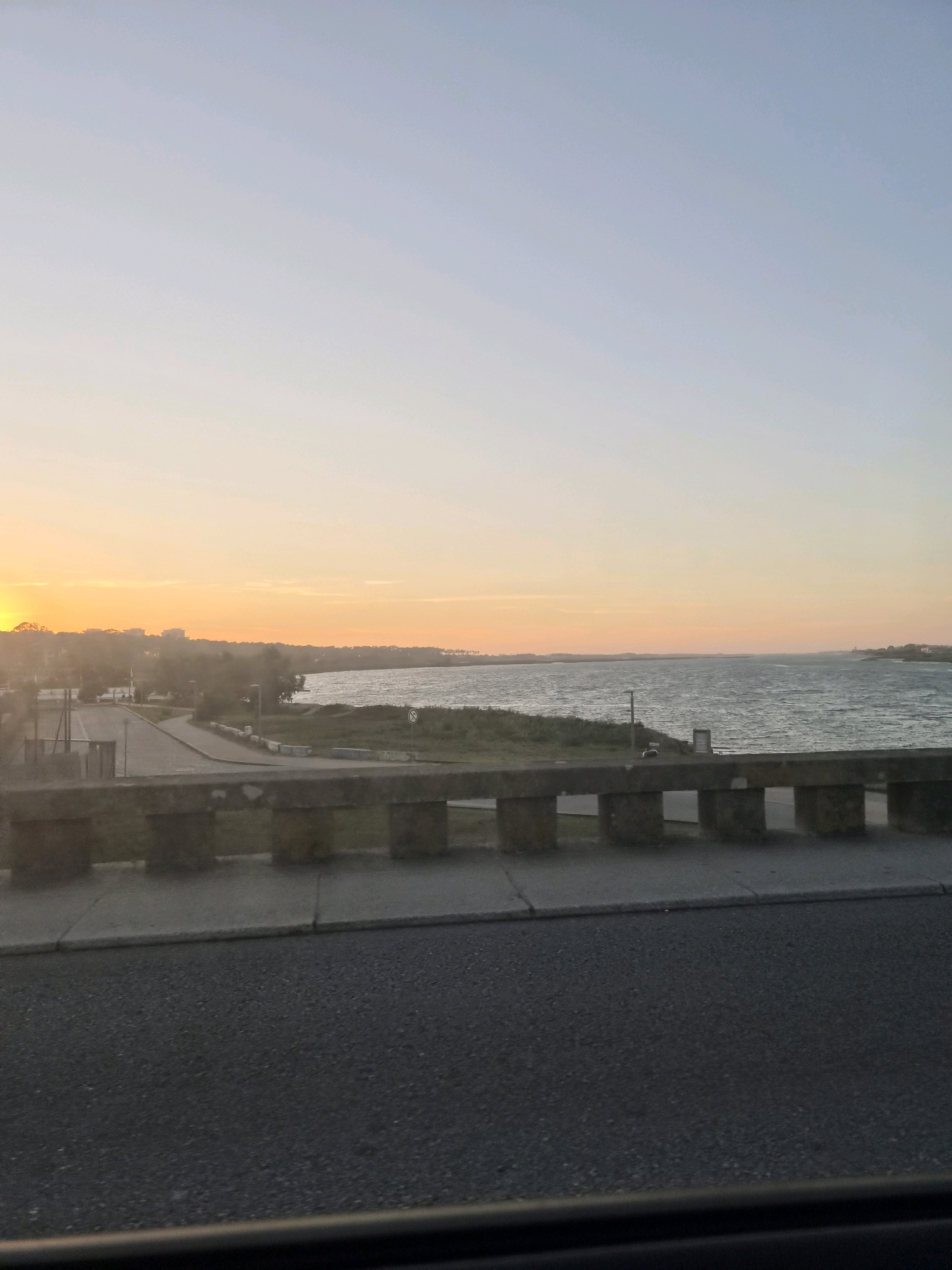 The image depicts a view from a bridge overlooking a body of water, likely the Cávado River estuary in Esposende, Portugal, during what appears to be sunset. The sky transitions from a soft orange glow near the horizon to a pale blue overhead. On the left, a road curves gently, bordered by low vegetation and some indistinct buildings in the distance. The water is rippled, suggesting a light breeze. A concrete railing with rectangular supports runs across the foreground, separating the roadway from the view. The roadway itself is asphalt and shows some texture. There are no people or vehicles clearly visible in the scene, and the overall atmosphere is calm and serene.