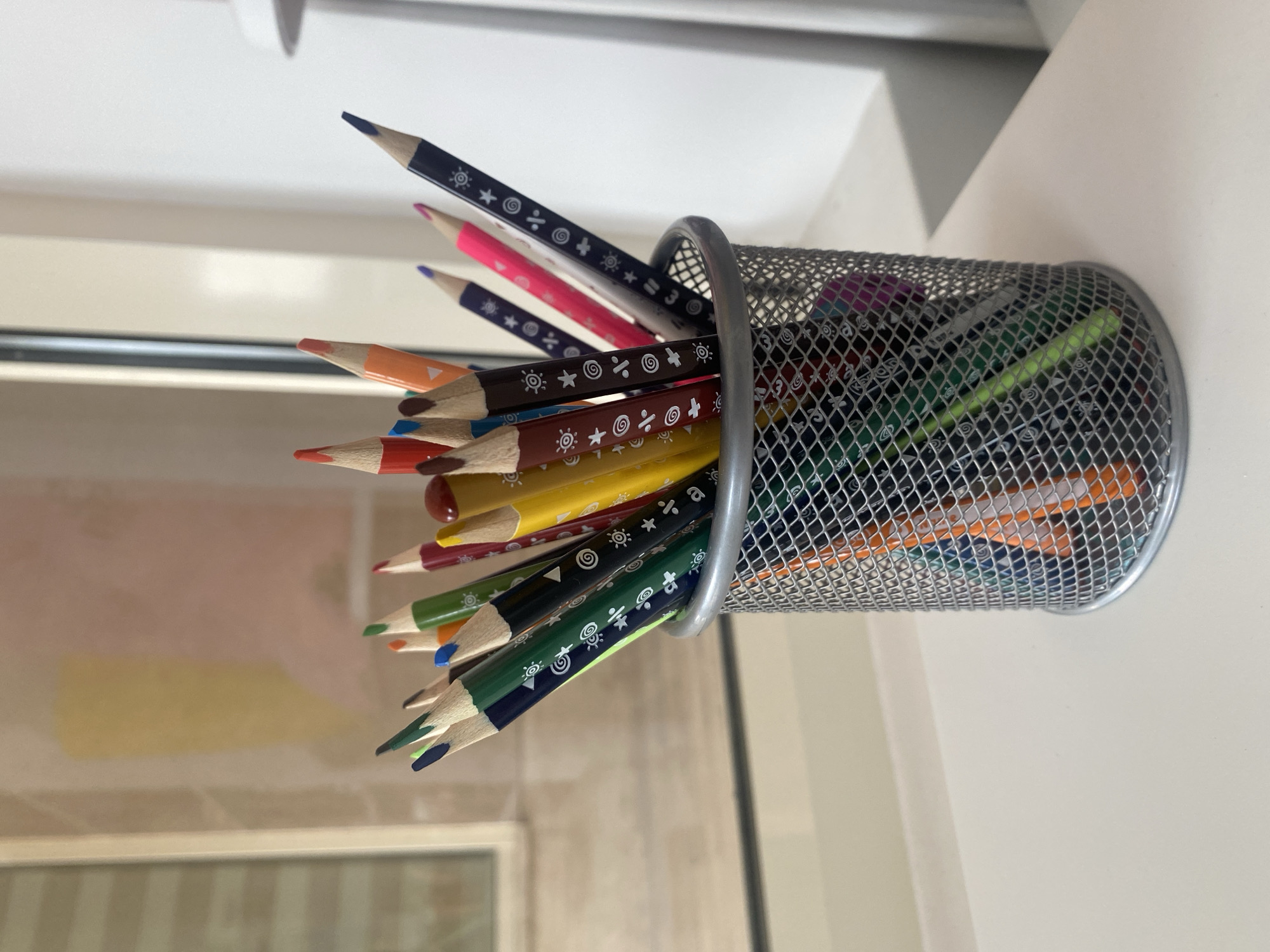 A wire mesh pencil holder, filled with colored pencils, sits on a white surface in an indoor setting. The pencils are arranged haphazardly, extending outwards from the container. The pencil holder is made of a shiny, silver-colored metal and appears to be cylindrical in shape. The pencils vary in color, including shades of blue, pink, orange, brown, yellow, and green, each pencil adorned with a white geometric pattern. The background consists of a white or light-colored wall, with vertical lines that seem to indicate a window frame. The scene is located in an Unknown city.