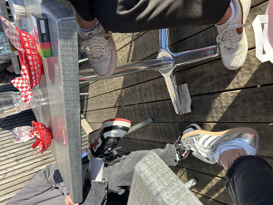 The image captures a high-angle view of the lower half of several individuals seated at an outdoor table, with their legs and feet visible. The setting appears to be a patio or deck, characterized by light brown wooden planks forming the ground surface. Bright sunlight casts strong, defined shadows across the wooden planks, suggesting a clear day.
In the upper right portion of the frame, one person's legs and feet are visible. This person is wearing dark, possibly black, pants and a pair of light-colored, likely white or off-white, Nike Air Force 1 sneakers. The Nike swoosh and heel tab on these shoes have a subtle reddish-brown or tan accent. White socks are also visible. Their feet are positioned near a shiny, chrome-plated metal base, which appears to be part of a table or chair support.
In the bottom left, another person is seated, wearing dark grey, cuffed jeans and high-top sneakers. These sneakers feature a black and white design with a prominent red star on the side and red detailing on the sole and heel. White socks are worn with these shoes.
Towards the bottom right, a third person's legs and feet are visible, also wearing dark grey or black cuffed jeans. This individual is wearing metallic silver sneakers with white stripes and laces, resembling an Adidas Samba style. White socks are visible, and a small, unidentifiable tattoo can be seen on the top of their right foot/ankle area.
The table itself is a grey, rectangular surface. On the table, several items are visible:
* A red plastic basket containing a red and white checkered paper liner is in the upper left.
* Another red plastic basket, holding crumpled white paper, is positioned further down the table.
* A black rectangular object, possibly an electronic device or vape, is on the table next to the first red basket.
* A small stack of colorful, rectangular items, possibly sticky notes, is next to the black object.
* A glass bottle or cup, and another clear glass, are also on the table.
* A white paper bag or container is on the ground near the foot of the person in the upper right.
* A crumpled white napkin or tissue is on the wooden planks near the chrome table/chair base.
The overall scene conveys a casual outdoor gathering, likely a meal or drinks, given the presence of the table and various items associated with dining.