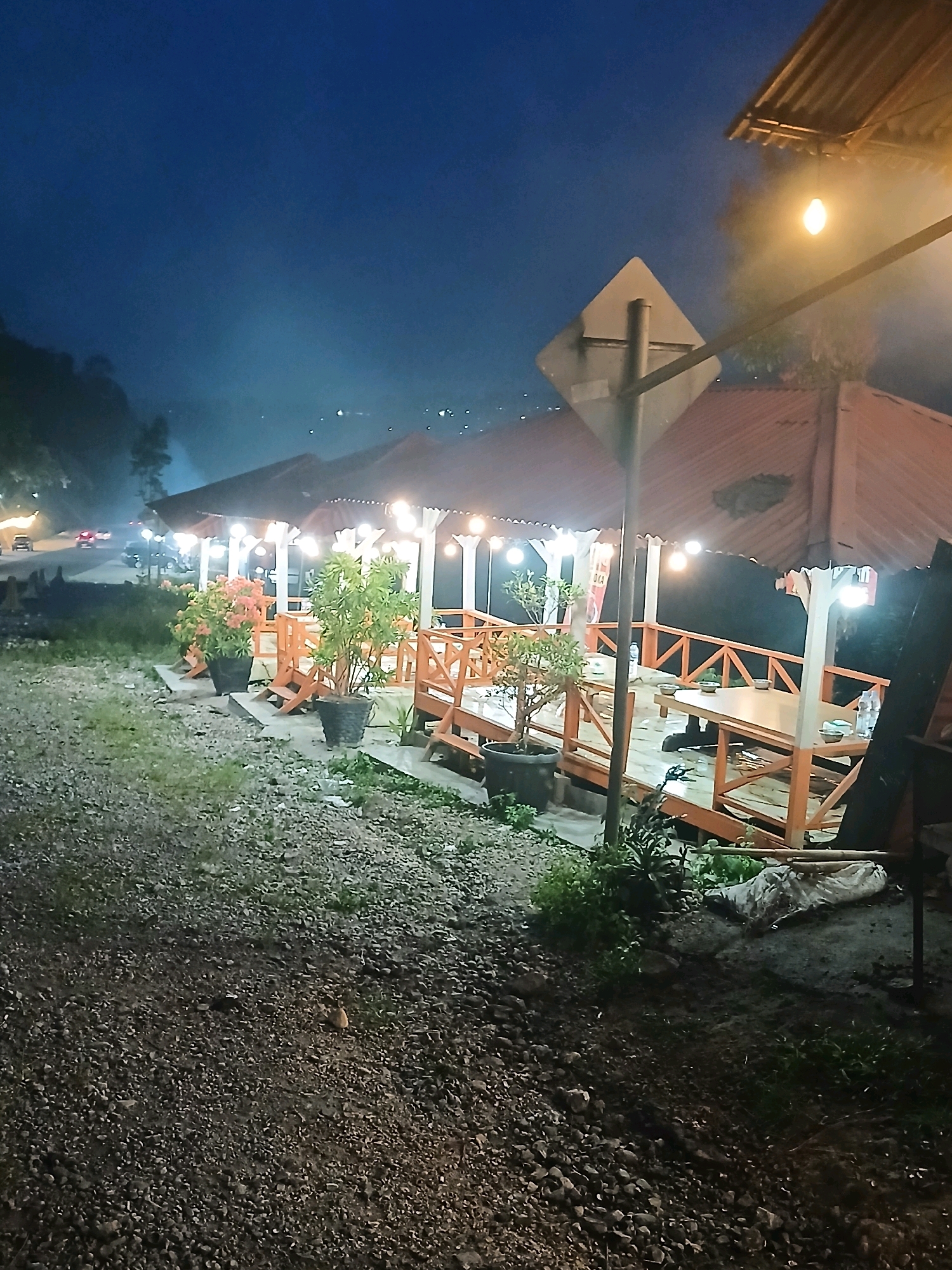 The image shows an outdoor seating area at dusk or night. Several covered gazebos with wooden railings and tables are visible. String lights are illuminated under the gazebos, and a single bare bulb hangs from a wire above a signpost. Potted plants, including one with orange flowers and others with green foliage, are placed near the seating areas. The ground in the foreground is covered with gravel, and a dirt path with some grass runs alongside it. In the background, there is a road with a few cars and distant lights, suggesting the presence of a town or city. The overall atmosphere is dimly lit and serene. The image captures an exterior scene, likely a restaurant or rest stop, situated in the Koto Gaek Guguak, Indonesia region. There are no people clearly visible in the image.