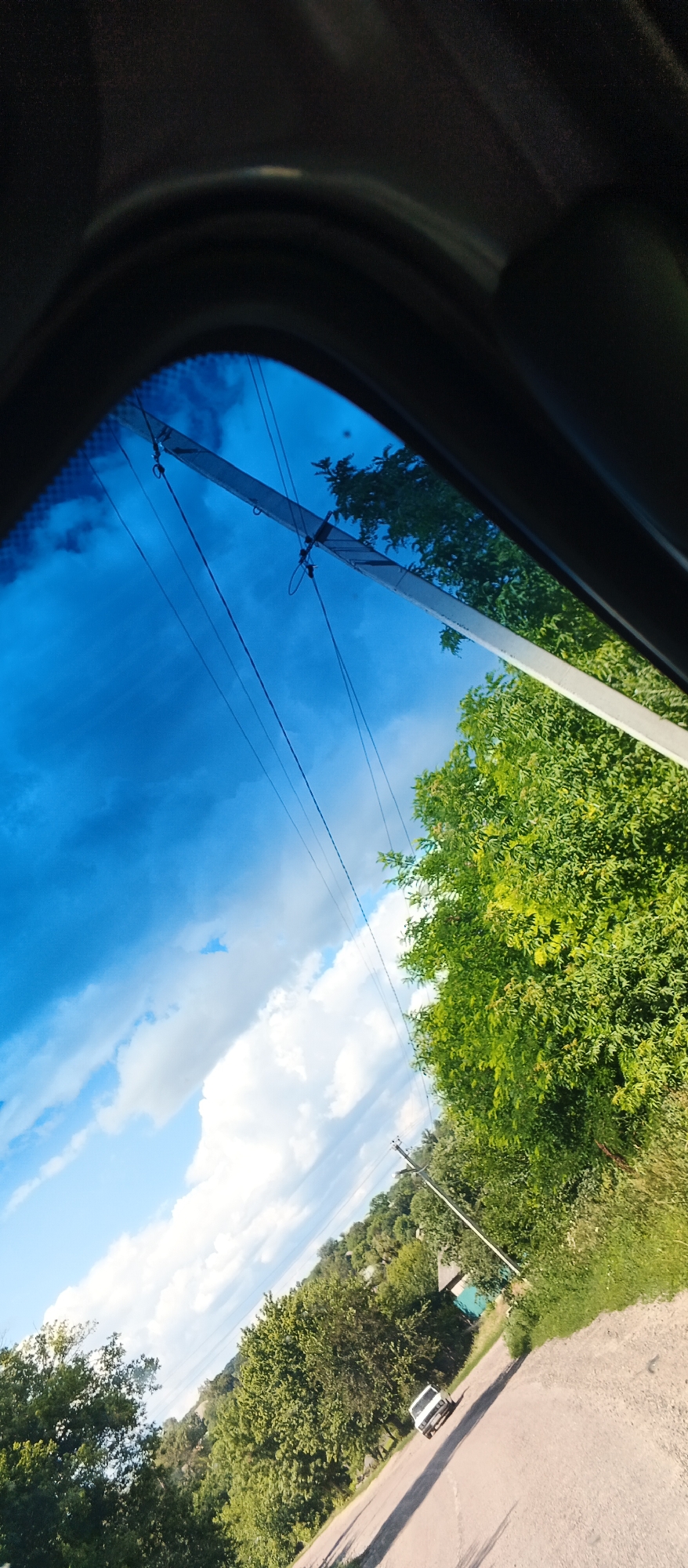 The image shows a view from inside a vehicle looking out through the windshield at a rural landscape. The sky is bright blue with scattered white clouds, and a utility pole with wires runs diagonally across the upper part of the frame. Lush green trees line the right side of the image, with more trees and vegetation visible in the distance. A narrow, paved road curves through the scene, and a white car is driving away from the viewer on this road. The overall impression is one of a sunny day in a countryside setting. The location is identified as Mala Smilianka, Ukraine.