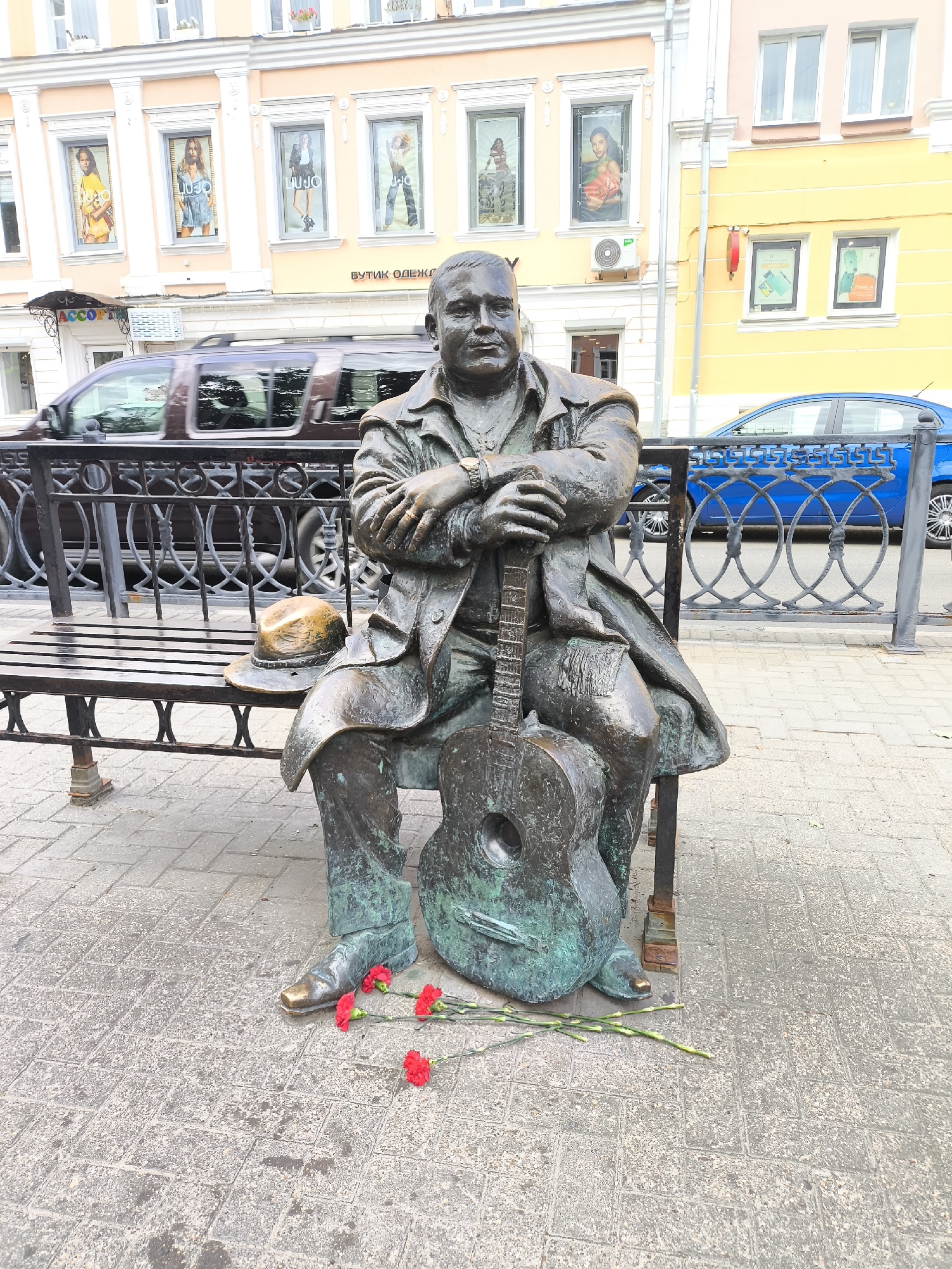 A bronze statue of a man sitting on a park bench holding a guitar is located on a sidewalk in the city of Tver, Russia. The statue is made of weathered bronze and depicts a man in a suit with his arms crossed over his guitar. A hat is placed on the bench beside him. In front of the statue, on the cobblestone ground, are three red carnations. Behind the statue, a dark-colored SUV is driving by, and a blue car is parked further down the street. The buildings in the background are brightly colored, with storefronts displaying advertisements. The scene appears to be a public space, possibly a square or a pedestrianized street, where the statue serves as a monument or art installation.