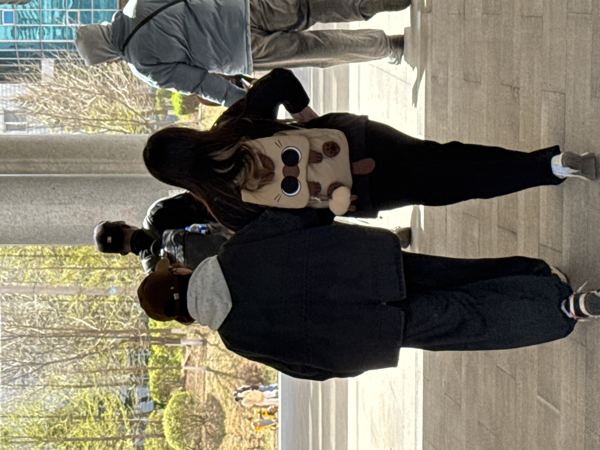 The image depicts a group of people walking outdoors on a paved surface that appears to be a sidewalk or plaza. The environment seems to be a public area with trees and potentially buildings in the background, suggested by the blurred reflections. The people in the image are dressed in casual clothing; some are wearing jackets, suggesting a moderate temperature. There is a person with a backpack that resembles an animal or cartoon character. The orientation of the image is sideways. The scene lacks elements allowing confirmation of the precise location in Zhongtun, China.