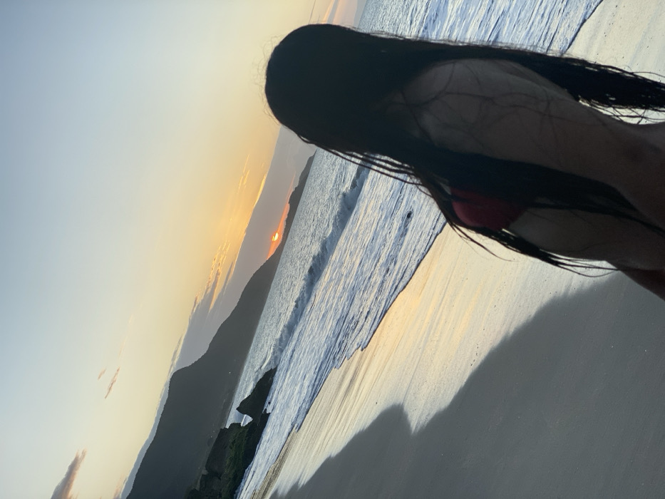 A silhouetted figure with long, dark hair stands on a sandy beach at sunset. The sun dips below the horizon, casting a warm glow on the water and sky. Gentle waves roll onto the shore, reflecting the light. A distant mountain range is visible against the colorful sky, adding depth to the scene. The figure's form suggests a bikini, and the composition emphasizes the natural beauty of the beach setting.