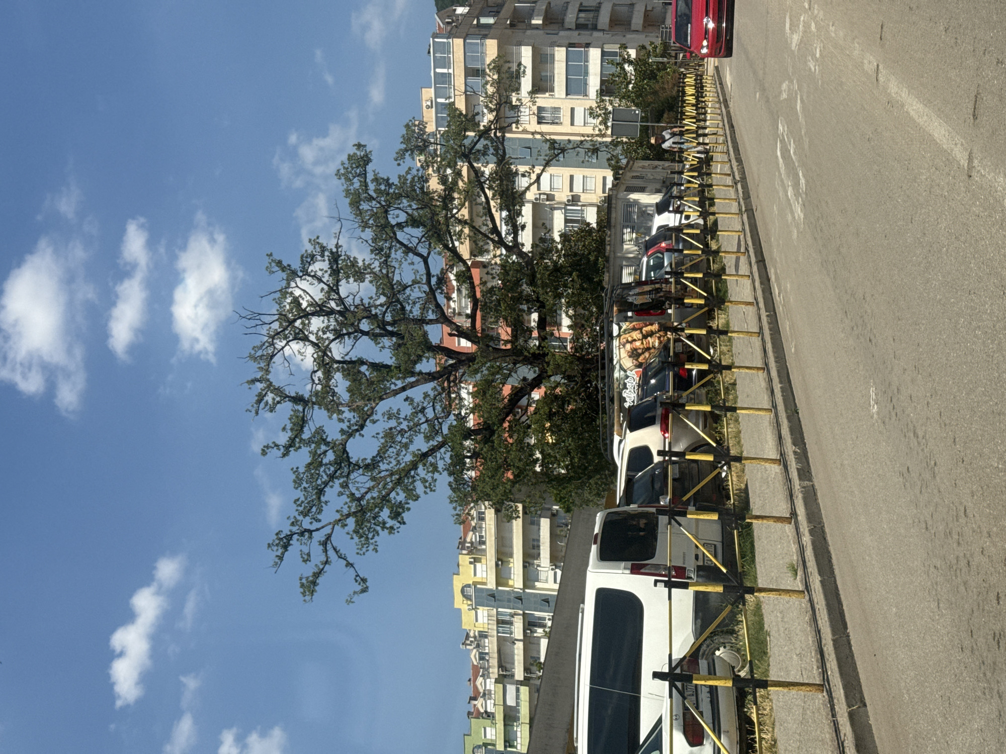 The image shows a street scene in Budva, Montenegro. On the left side of the street, several vehicles are parked, including a white van and a red car. A yellow and black striped barrier separates the parking area from the sidewalk. In the background, there are several multi-story buildings with colorful facades. A large tree with sparse foliage stands in front of the buildings. The sky is blue with scattered white clouds. A person is walking on the sidewalk. The overall scene depicts a typical urban environment on a sunny day.