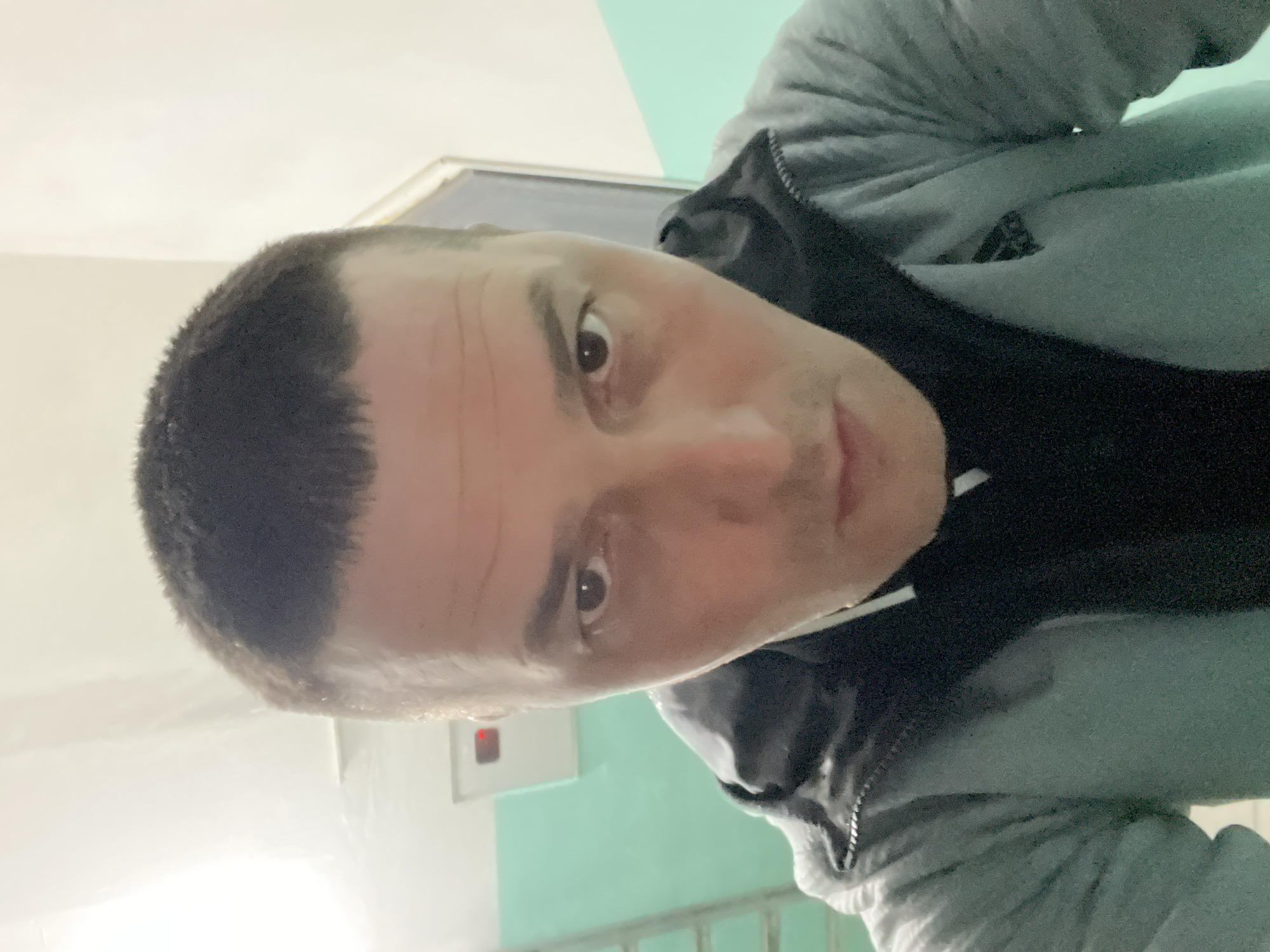 A close-up, slightly tilted, selfie of a man with short dark hair and light skin, wearing a black hoodie under a gray zip-up sweatshirt. He is looking directly at the camera with a neutral expression. The background consists of a white and turquoise painted wall. A white frame can be seen above the turquoise section of the wall. A small red object is partially visible on the white wall. Below the turquoise section of the wall, a gray or silver railing is present. The location is Unknown, Unknown.