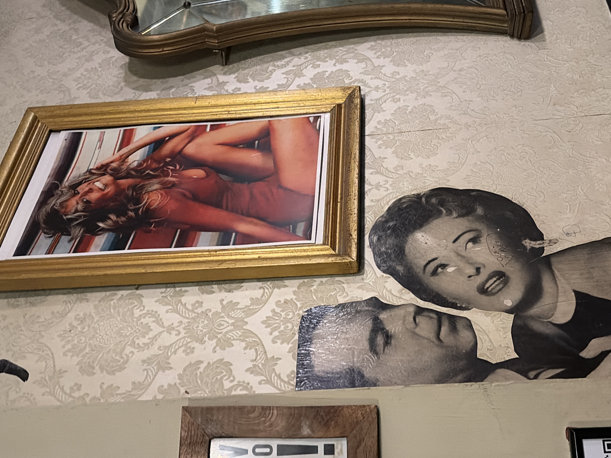 The image shows a close-up of a wall adorned with a damask-patterned wallpaper. Mounted on the wall are three framed items. On the left, a framed picture with a wooden frame contains a white background with black lettering that reads "V" and "0!". To the right of this, a framed photograph in a gold frame depicts a woman in a red swimsuit. To the far right, a vintage-style ornate gold mirror is partially visible. Below the framed photograph of the woman in the red swimsuit, a collage of two black and white photographs is attached directly to the wallpaper. One photograph shows the head and shoulders of a man, and another, overlapping and to the right, shows the head and shoulders of a woman with a distressed expression and what appears to be writing on her cheek. The scene is indoors.