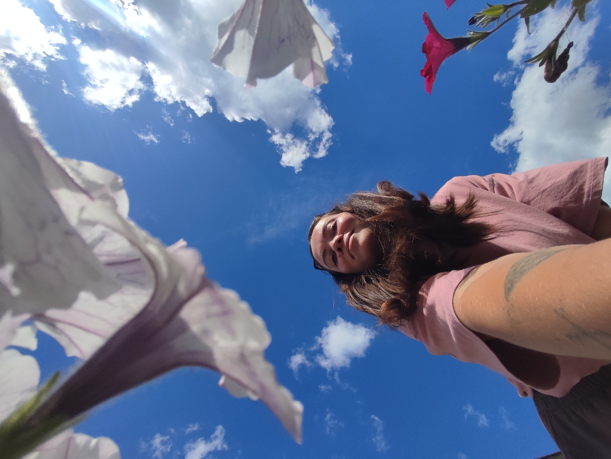 This is a low-angle, wide-shot, eye-level, selfie-style photograph taken outdoors under a bright blue sky with scattered white clouds. In the foreground, large white and purple petunia flowers are in focus, with a single pink petunia visible in the upper right quadrant of the image. A young person with long, dark brown hair is looking down towards the camera, their face partially obscured by shadow. They are wearing a light pink t-shirt and dark pants, and have a visible tattoo on their upper arm. The camera appears to be positioned at ground level, looking up. The overall impression is one of a sunny, pleasant day, possibly in a garden or park, with the subject interacting playfully with their surroundings. The lighting suggests it is daytime, likely late morning or afternoon. There is no visible text in the image.