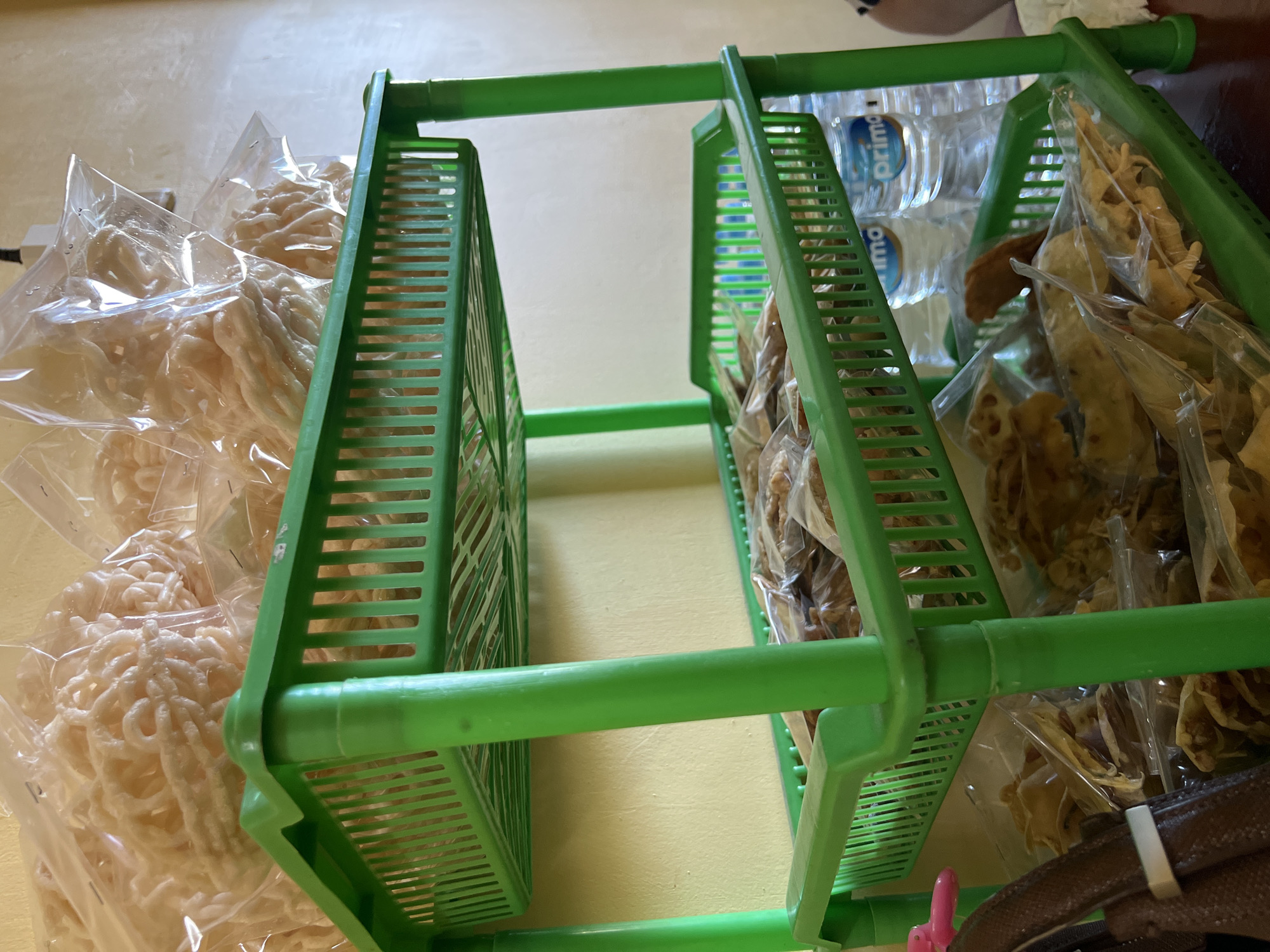 The image shows a green, three-tiered plastic shelving unit displaying various packaged snacks. The top shelf holds large, clear plastic bags filled with pale, coiled snacks, possibly crackers or krupuk. The second shelf contains clear plastic bags filled with smaller, irregularly shaped, golden-brown snacks. The bottom shelf also holds clear plastic bags of similar golden-brown snacks, with two bottles of "Prima" bottled water visible behind them. The shelving unit is placed against a textured, yellow wall. To the left of the unit, a portion of a dark brown bag or strap with a white tie is visible. The scene appears to be indoors, likely a small shop or stall, given the presentation of goods. The context suggests a location in Gianyar, Indonesia, where such snacks are commonly sold.