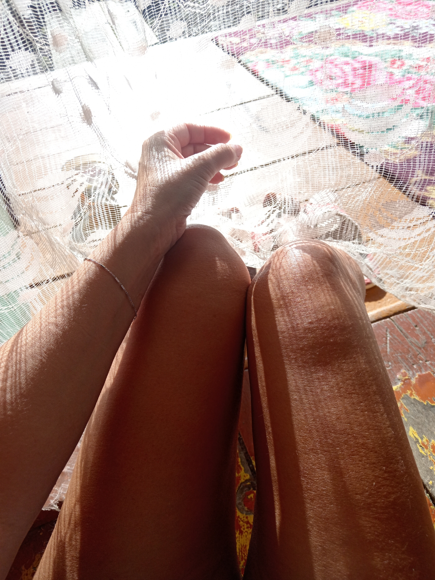 An overhead shot captures a person's legs and arm resting on a wooden surface covered by a white lace-like fabric with a floral patterned textile underneath. Sunlight casts a striped shadow across the fabric and skin. The person is wearing a delicate silver bracelet on their left wrist. The image does not contain enough information to determine the location in Novopokrovka, Russia.