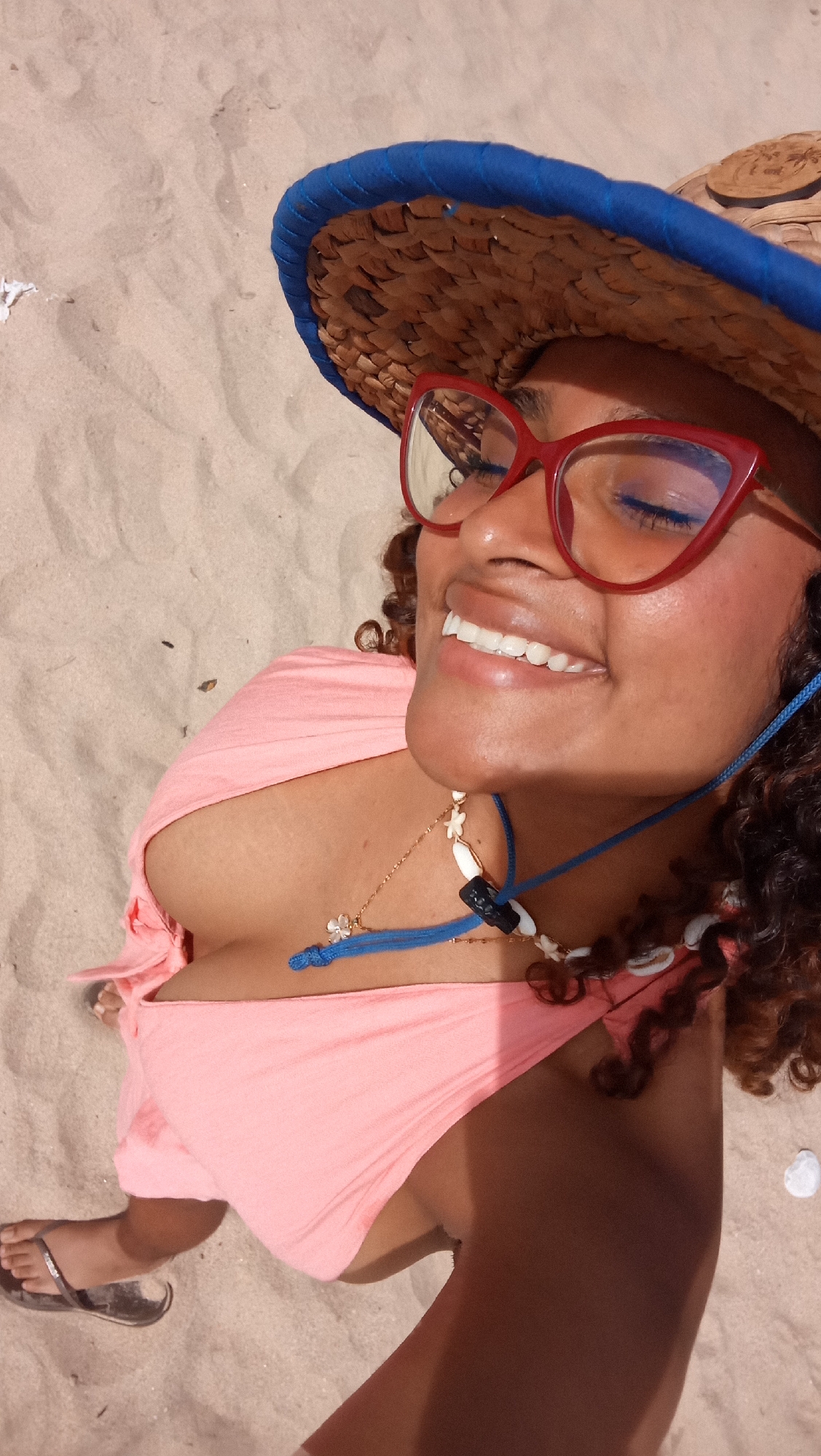 A woman is lying on the sand in El Tunal, Venezuela, wearing a pink bikini top, a straw hat with a blue trim, and red-framed glasses. She is smiling with her eyes closed. A necklace with shells and a blue cord is around her neck. Her feet, wearing flip-flops, are visible on the left side of the frame. The sandy environment suggests a beach or coastal area.