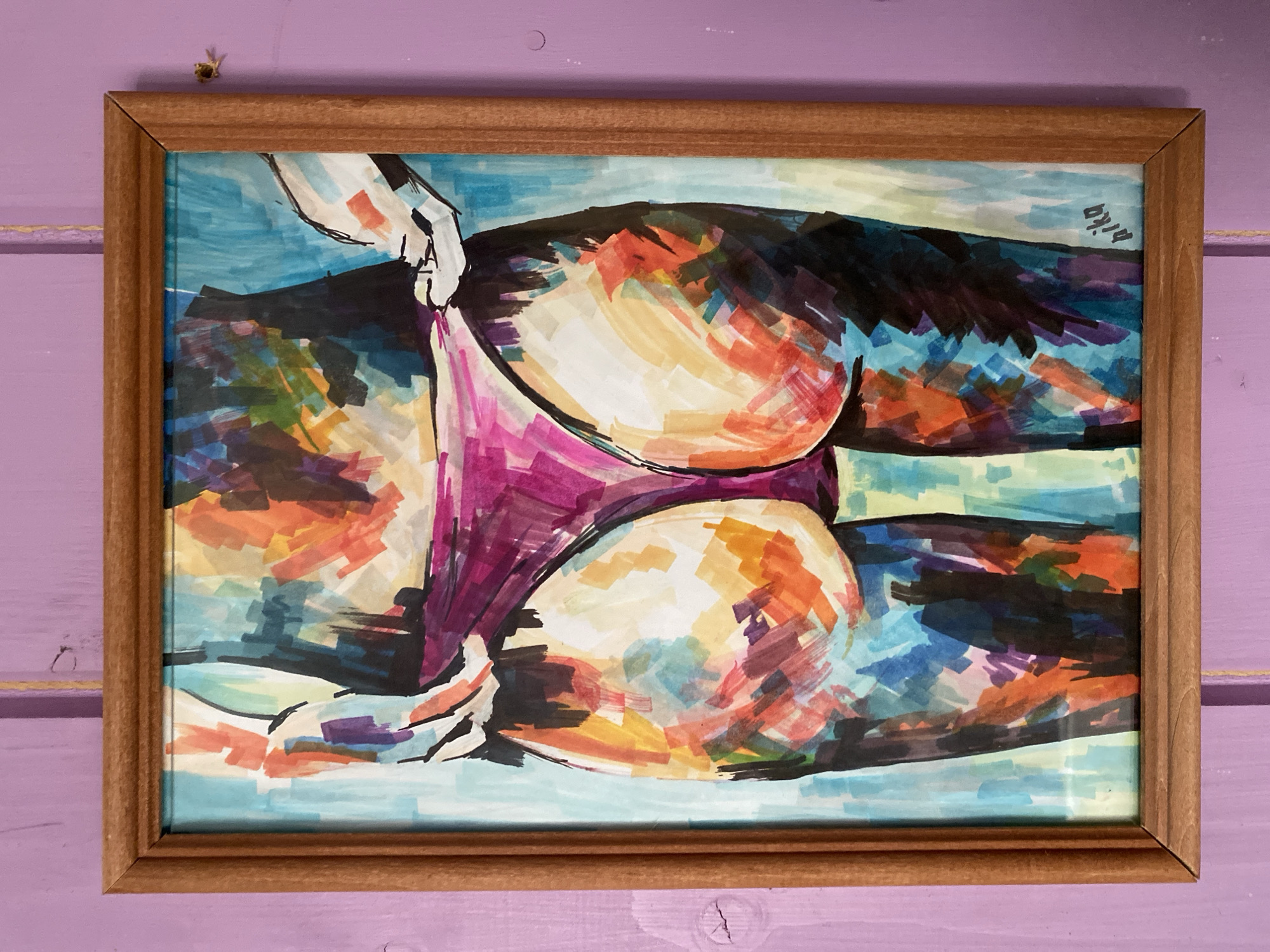 The image is a framed artwork of a person's back and buttocks wearing purple underwear. The artwork is abstract, rendered in bold strokes of various colors, including blues, purples, oranges, and yellows. The frame is a simple wooden one. The artwork is hanging on a wall that is painted a light purple color and appears to be made of vertical wooden planks. The lighting suggests it is daytime, with a slight shadow cast to the right of the artwork. A small insect is visible on the wall to the right of the frame. The location is not specified in the image.