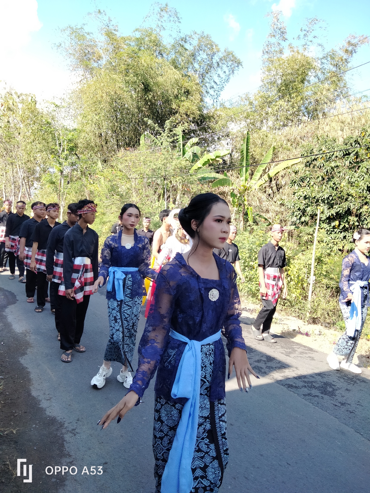 In Bendorejo, Indonesia, a group of young people, some in traditional attire, walk along a paved road bordered by lush vegetation. In the foreground, a woman in a blue lace kebaya and patterned skirt, with her hair styled up, walks forward, her hand extended. To her right, another woman in a similar blue outfit walks with a white sash. Behind them, a line of young men in black shirts and checkered sashes walk in single file. The background features a dense arrangement of trees and bamboo, under a bright blue sky with scattered clouds. The scene appears to be a procession or cultural event.