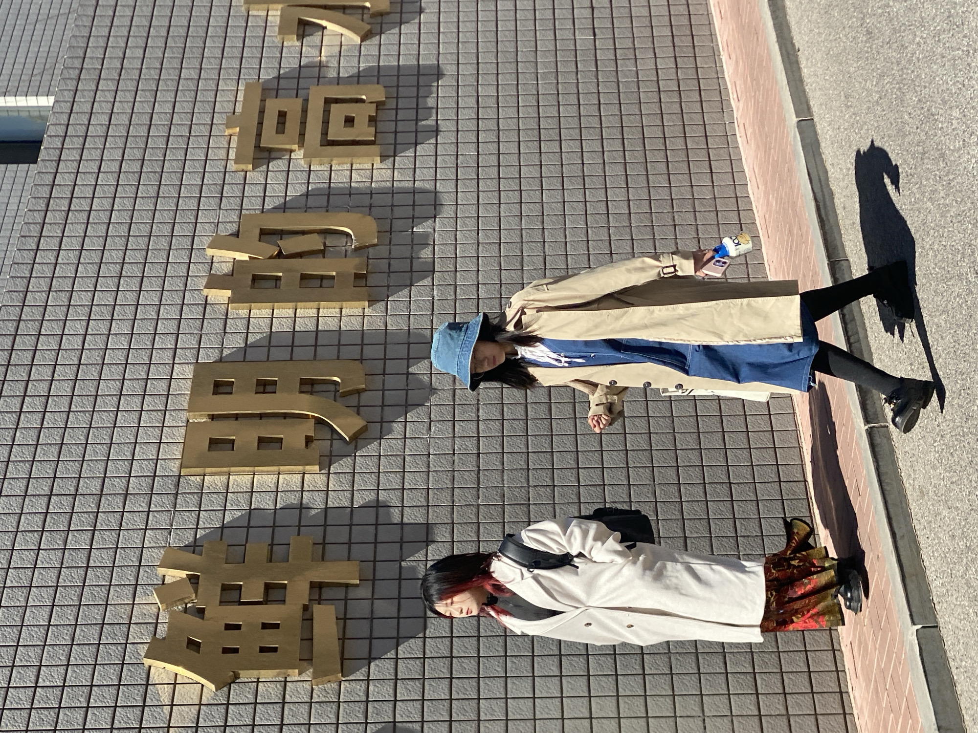 The scene depicts two individuals on a sunny day in the city of Zhongtun, China, situated on a red brick sidewalk alongside a building. The building's facade is composed of grey square tiles, adorned with large, gold-colored Chinese characters that read '鲜明的力量' (Vivid Power), casting shadows on the wall. The individual on the left stands with their left side towards the viewer, facing left, with dark hair featuring red highlights. They wear a white long coat over a black garment, a long patterned skirt with red, green, and gold tones, and black chunky shoes, carrying a black backpack on their left shoulder. The individual on the right is in motion, walking leftwards, attired in a blue bucket hat, a tan trench coat, a blue denim dress or overalls, black leggings, and black chunky shoes. This person holds a white smartphone and a small white bottle with a yellow cap and label in their right hand. The sidewalk is bordered by a grey curb and an asphalt street.